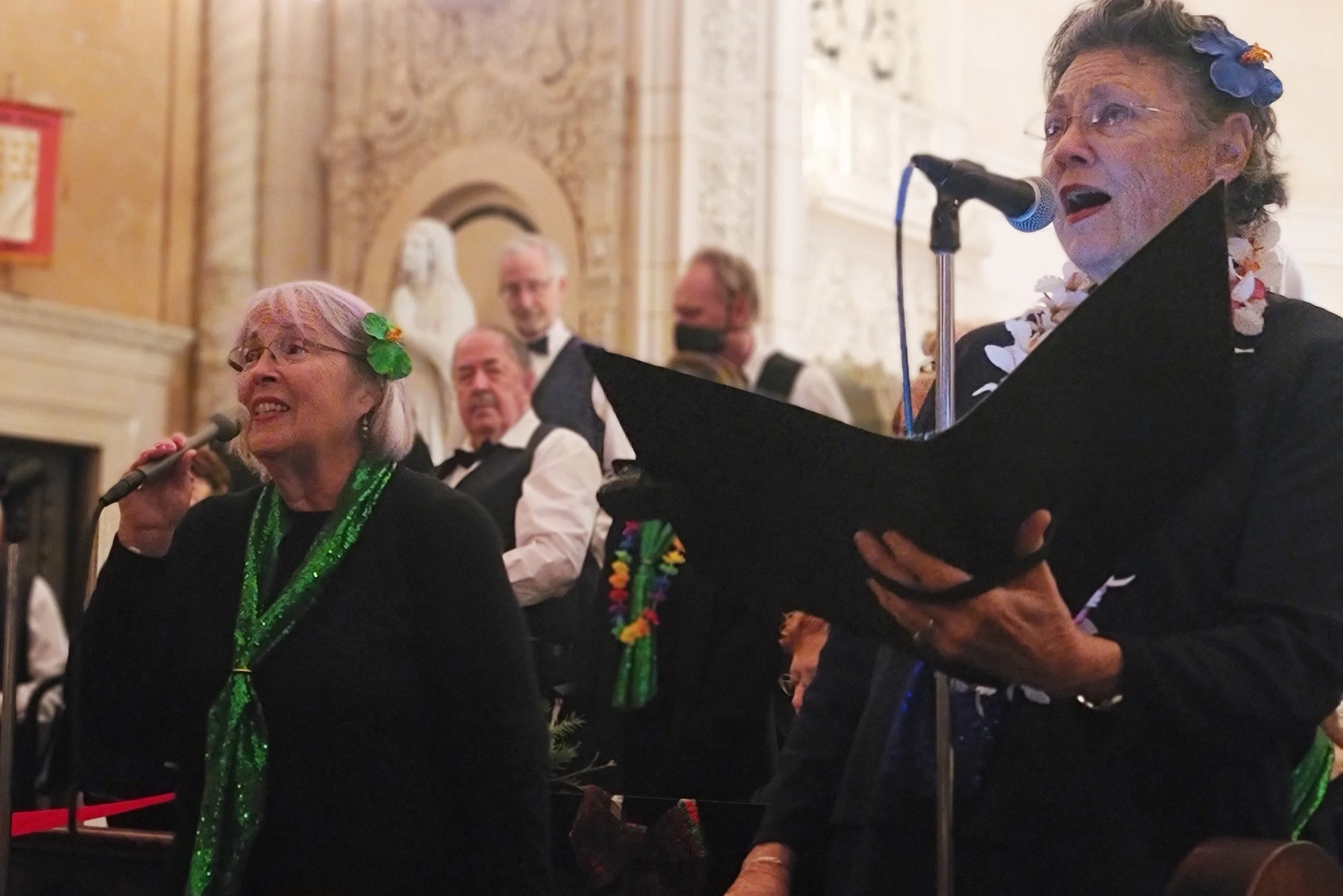 Two elderly women singing into microphones at a formal event, wearing floral accessories and leis, with a group of men in tuxedos and bow ties in the background.