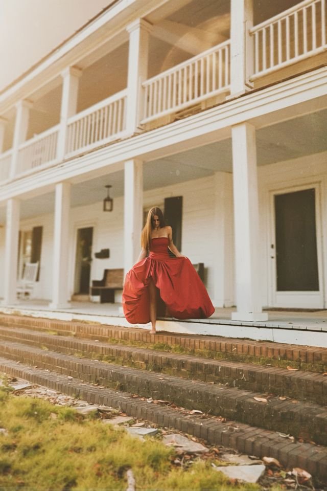A woman in a strapless red gown walking down the steps of a white house with a porch and balconies.