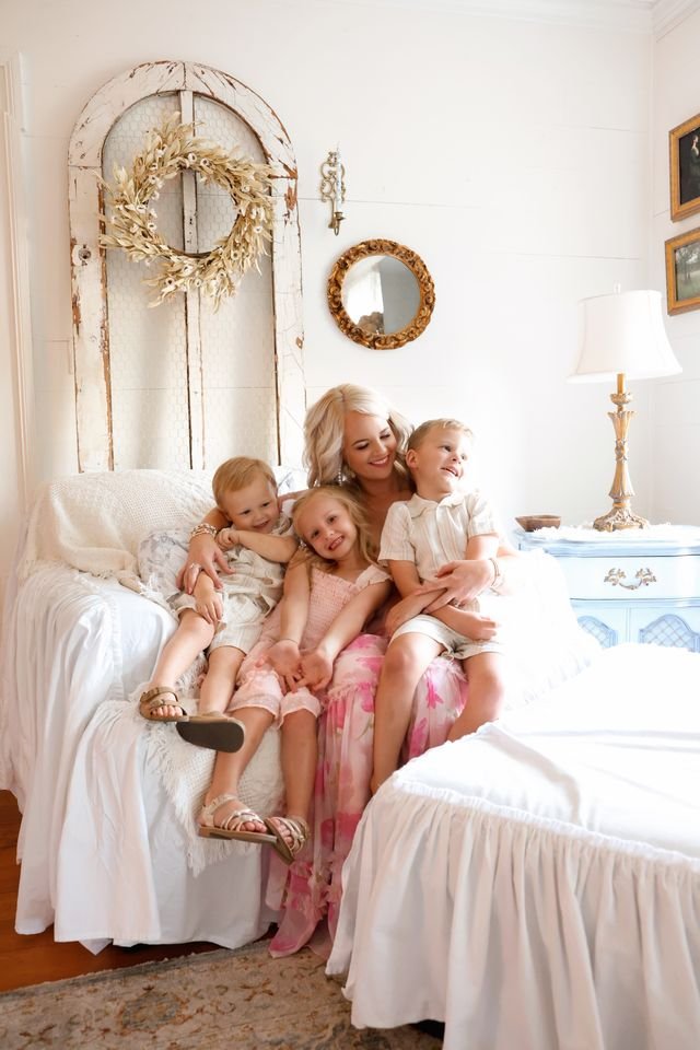 A woman sitting on a white sofa with four children, laughing and hugging each other in a bright, cozy living room with vintage decor and a white bedding on a nearby bed.