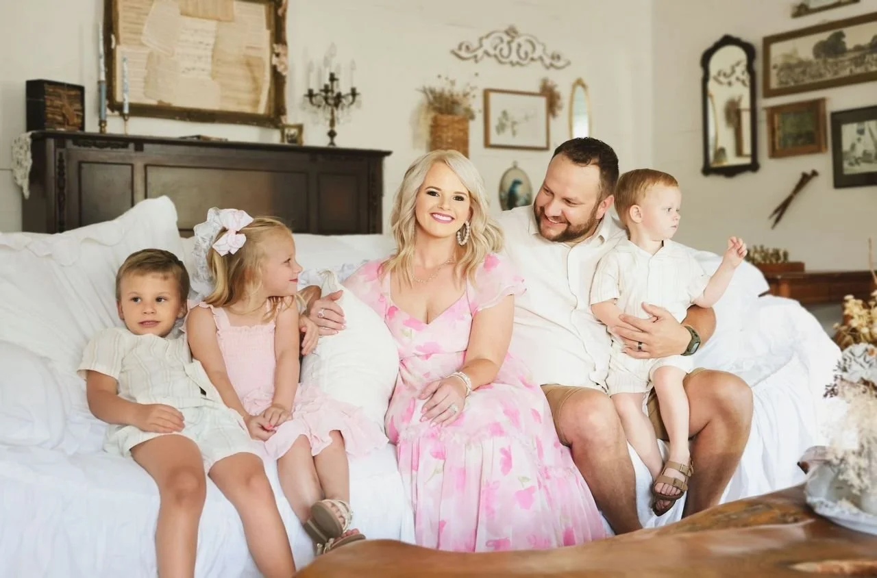 Family sitting on a white sofa in a decorated living room, including a woman in a pink dress, a man, and three young children.
