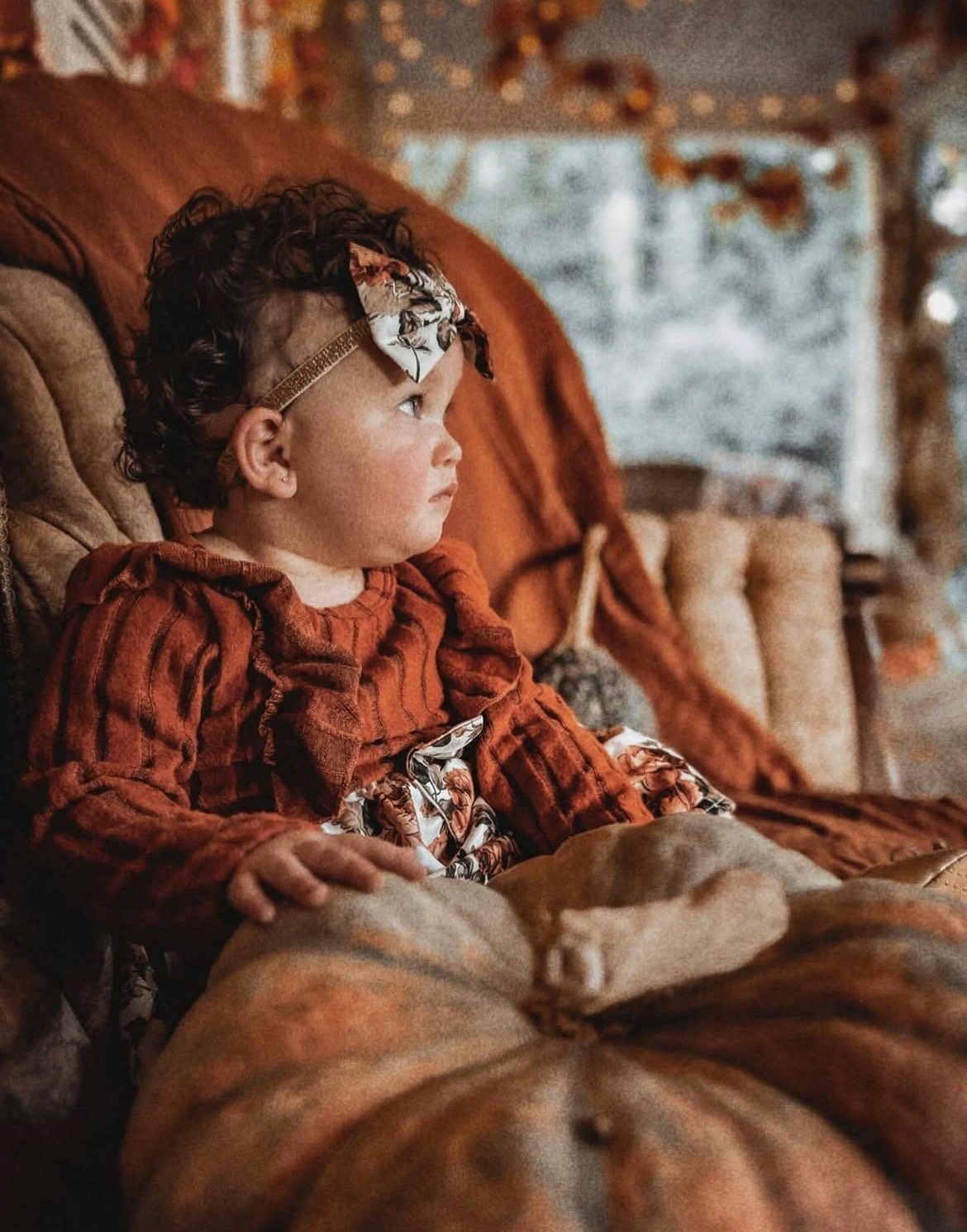A young girl sitting on a vintage sofa in a cozy, warmly lit room with a window and autumn leaves visible outside. She has curly hair, a headband with a bow, and is wearing a rust-colored dress. She is looking attentively to the side, with a calm exp
