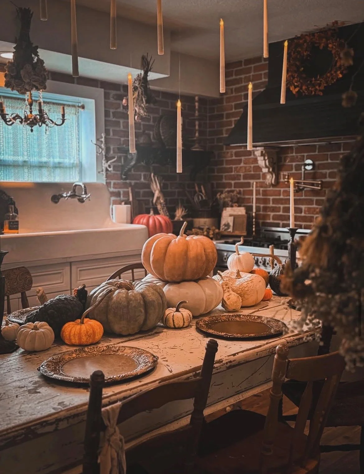 A rustic dining room decorated for fall with pumpkins on a distressed wooden table, surrounded by vintage chairs, with candles hanging from the ceiling and a brick wall in the background.