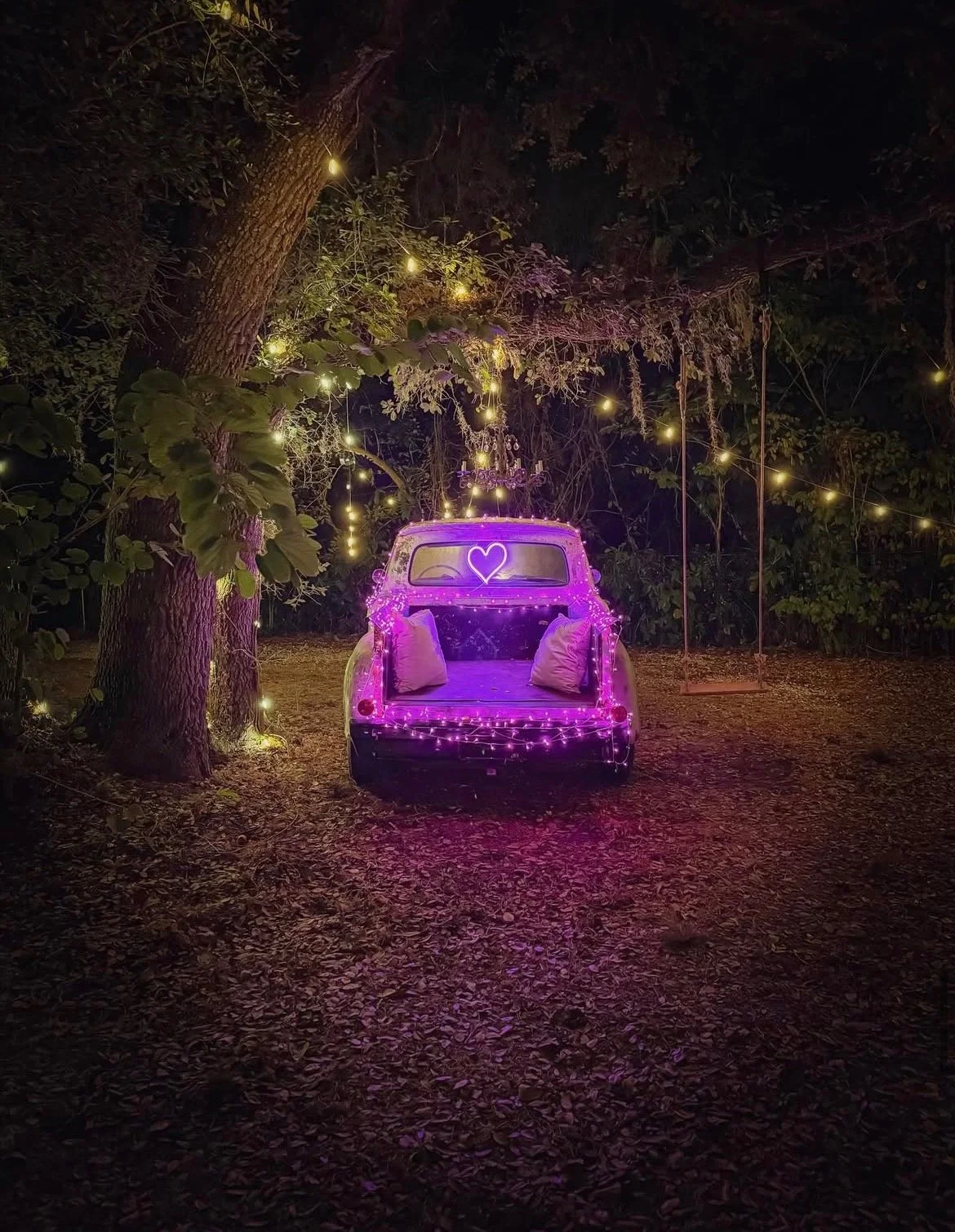 An outdoor scene at night featuring a vintage car decorated with pink and purple fairy lights, with a neon purple heart sign on the back window, surrounded by trees and hanging string lights, creating a romantic ambiance.