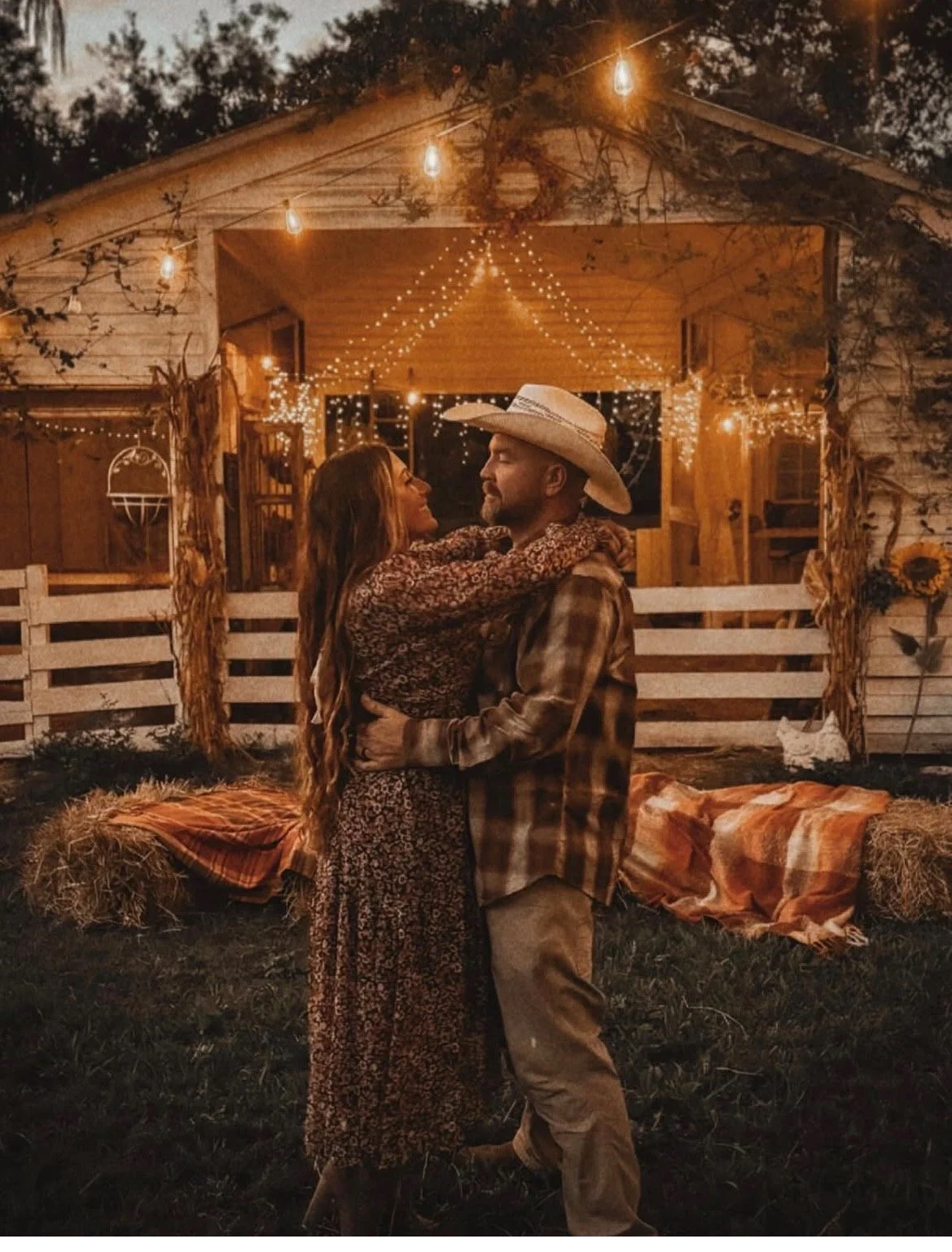 A romantic couple dancing in front of a decorated barn at night, illuminated by string lights and hanging bulbs, with hay bales and rustic decorations in the background.