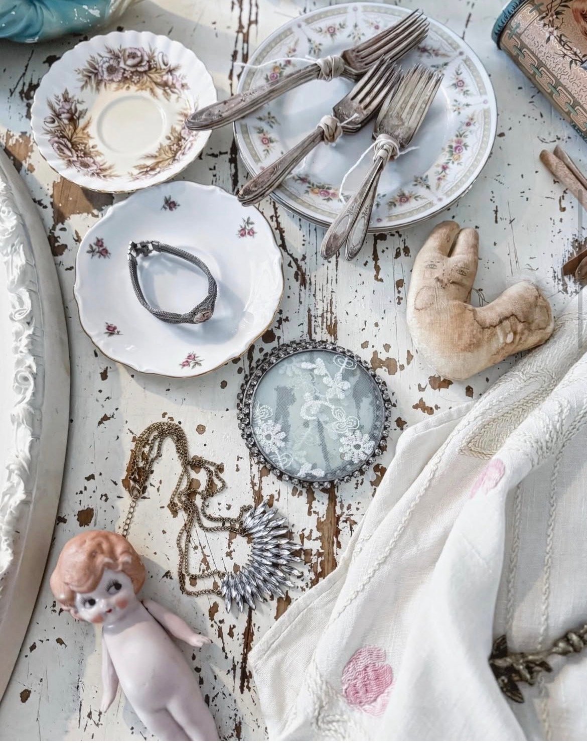 Collection of vintage jewelry, decorative plates, a small porcelain angel figurine, and assorted cutlery on a distressed white wooden surface.