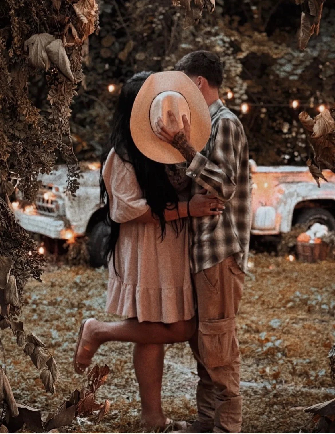 A couple stands close together, with the woman kneeling on one knee, holding a straw hat in front of the man's face. The woman has long black hair and tattoos on her arm, while the man has short dark hair and is wearing a plaid shirt and cargo pants.