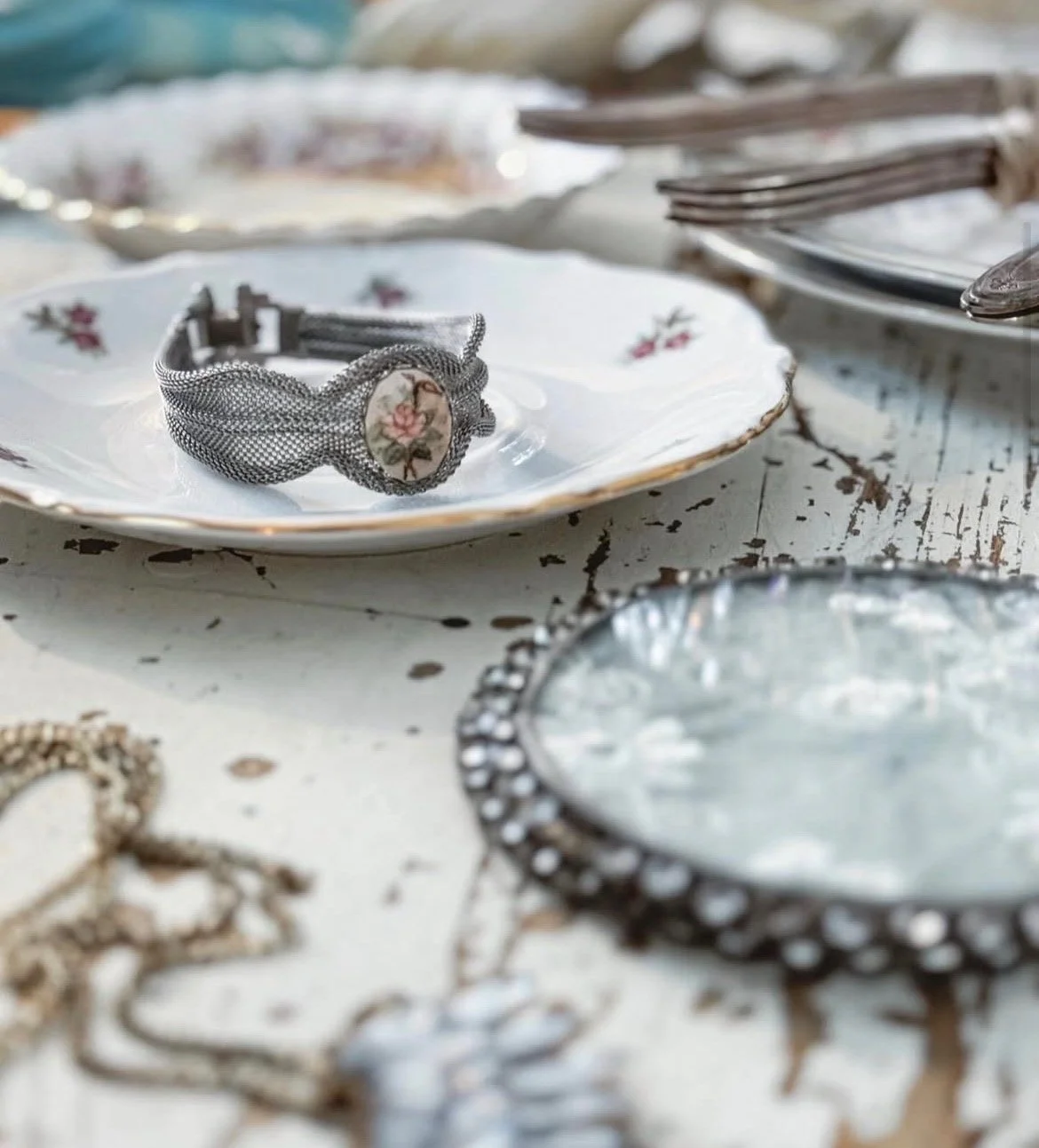 A vintage silver bracelet with a porcelain floral cameo is placed on a white floral patterned plate, surrounded by other jewelry and decorative items on a distressed wooden surface.