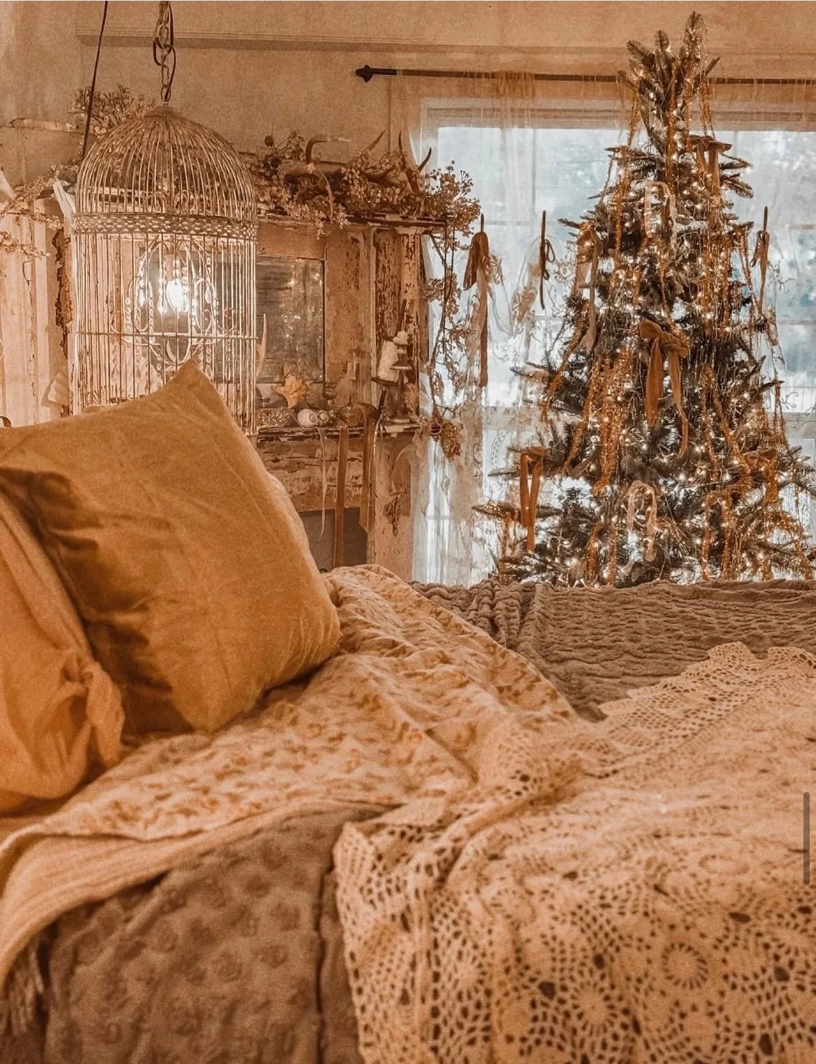 A cozy living room decorated for Christmas with a tree adorned with lights, bows, and ornaments. There is a large birdcage, vintage decor, and a bed with blankets and pillows in the foreground.
