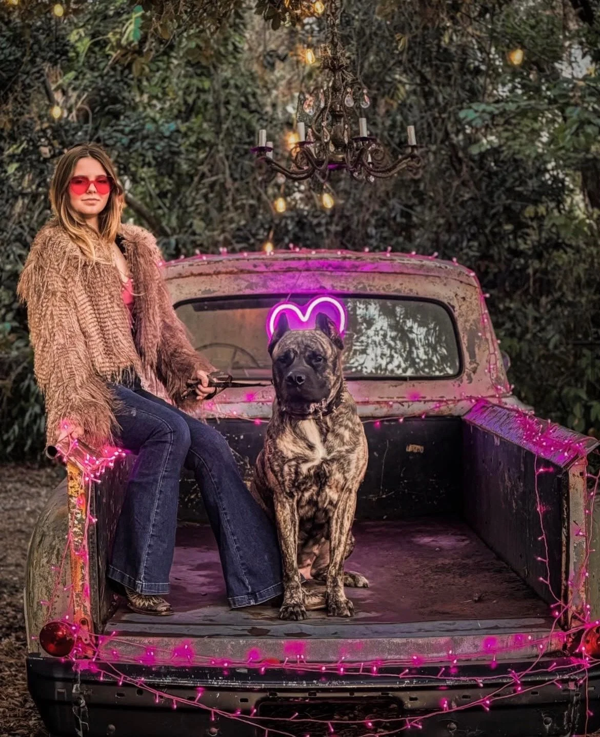 A woman with brown hair wearing a brown fur coat and red sunglasses sitting on the edge of an old, rusty truck bed decorated with pink string lights. A brindle pitbull sitting beside her. Behind them, a neon pink heart sign and a chandelier hang abov