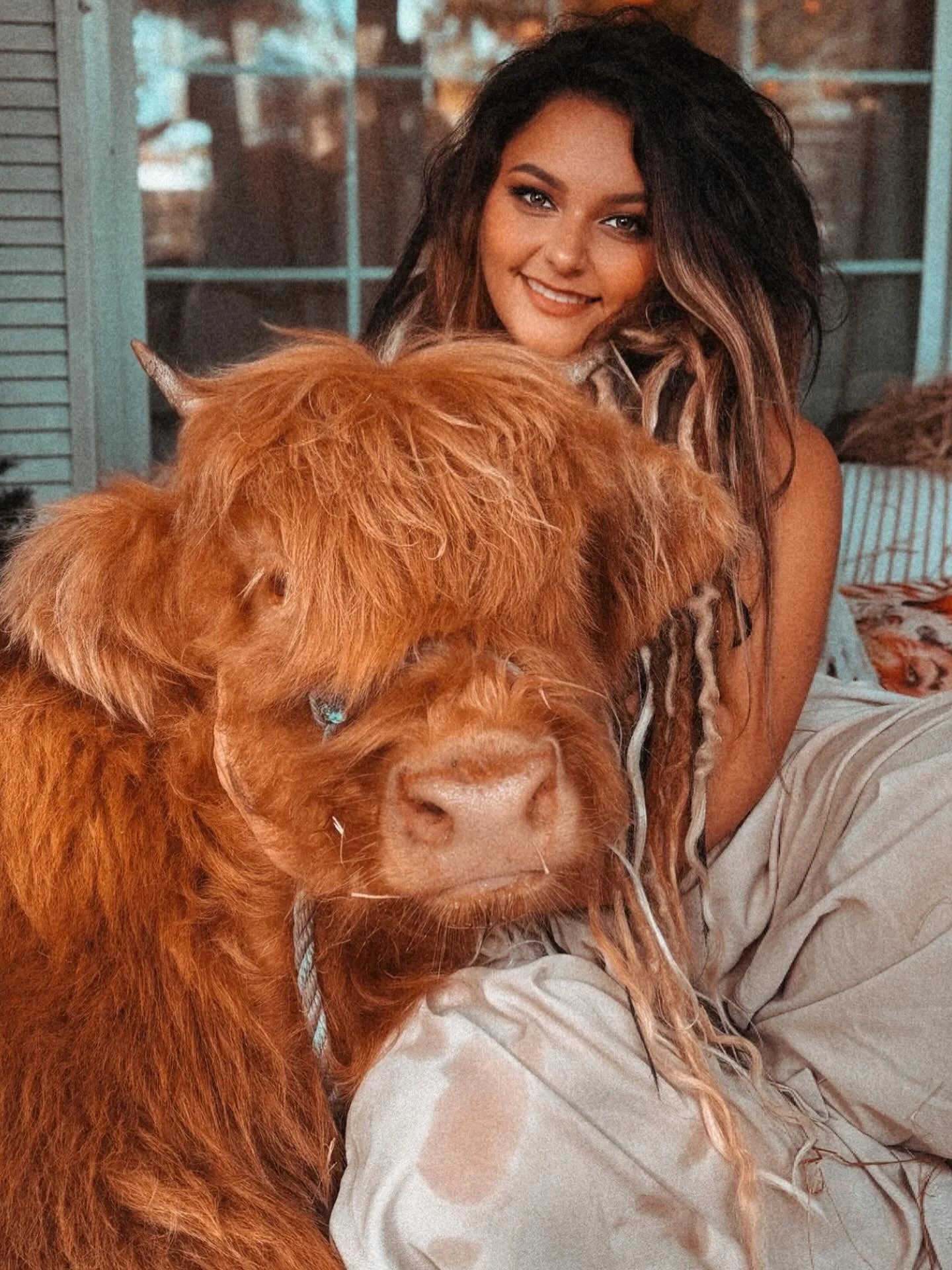 Woman with long, dark hair smiling while cuddling a large, fluffy, orange-colored dog with a thick coat, indoors near a window.