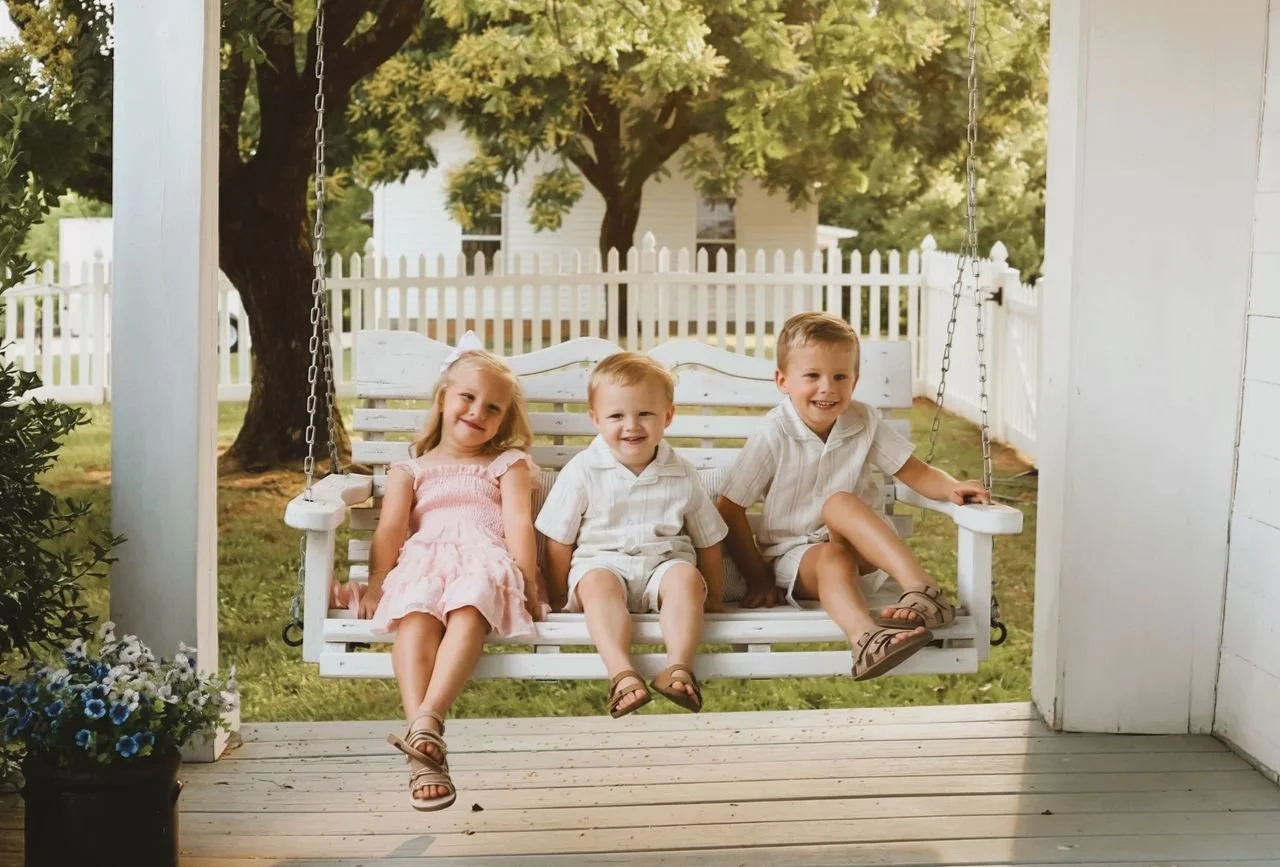 Three children sitting on a porch swing, smiling, with a white picket fence, trees, and a house in the background.