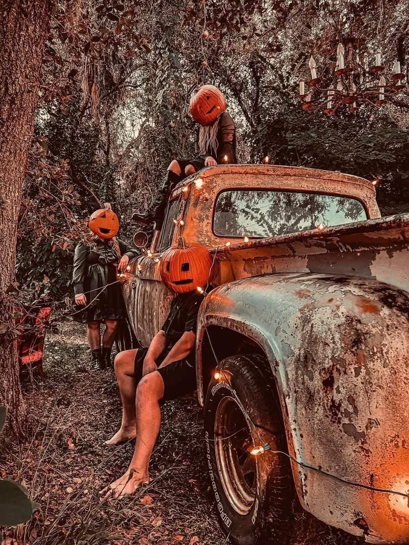 Four people with pumpkin masks in a wooded area decorated with Halloween lights around an old rusty pickup truck.