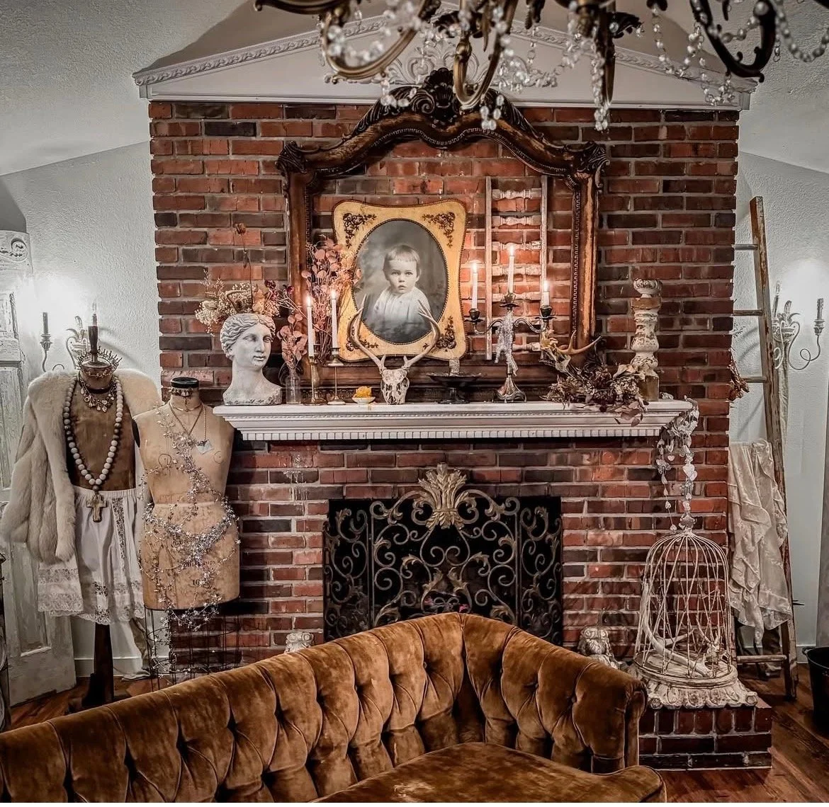 Decorative vintage living room with a brick fireplace, ornate mirrors, an antique portrait of a child, candles, jewelry mannequins, and vintage furnishings including a velvet sofa and birdcage.