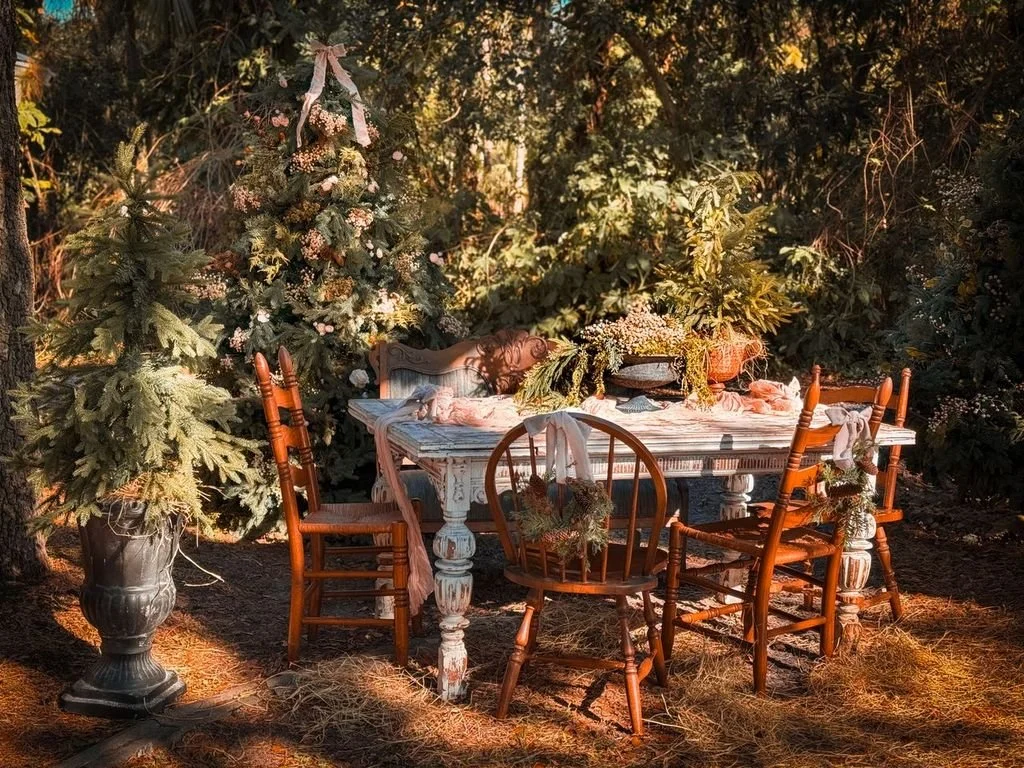 A vintage-style outdoor table surrounded by wooden chairs in a forested setting with Christmas trees and greenery for decoration.