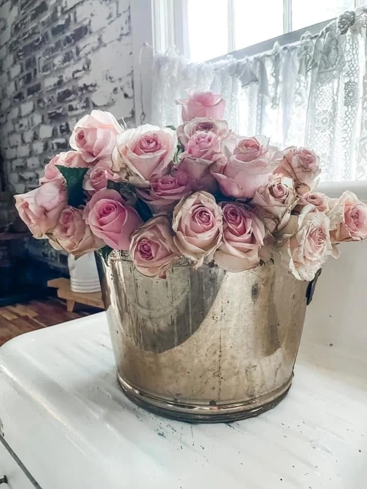 A silver metal bucket filled with pink and cream roses sits on a white surface in a room with a window, lace curtains, and a brick wall.