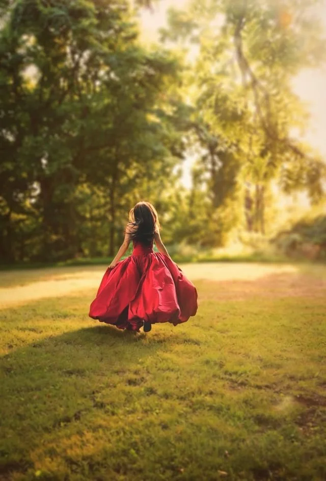 A girl in a flowing red dress walking away in a sunlit park with green trees and grass.