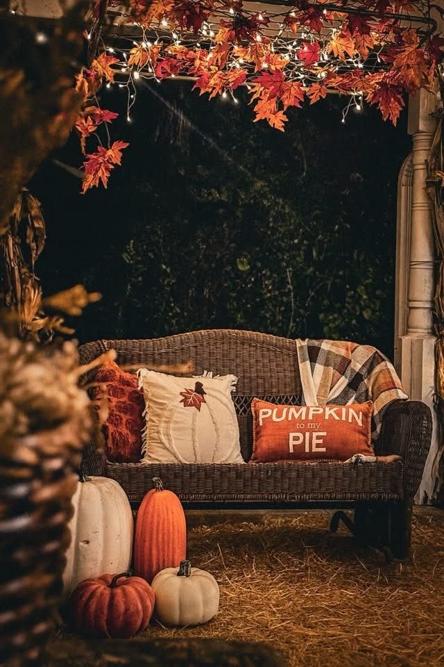 A porch decorated for fall with a wicker bench, autumn pumpkins on the ground, and cushions with fall-themed designs, including a pillow that says 'Pumpkin to my Pie'. Hanging red and orange leaves and string lights add to the cozy autumn atmosphere.