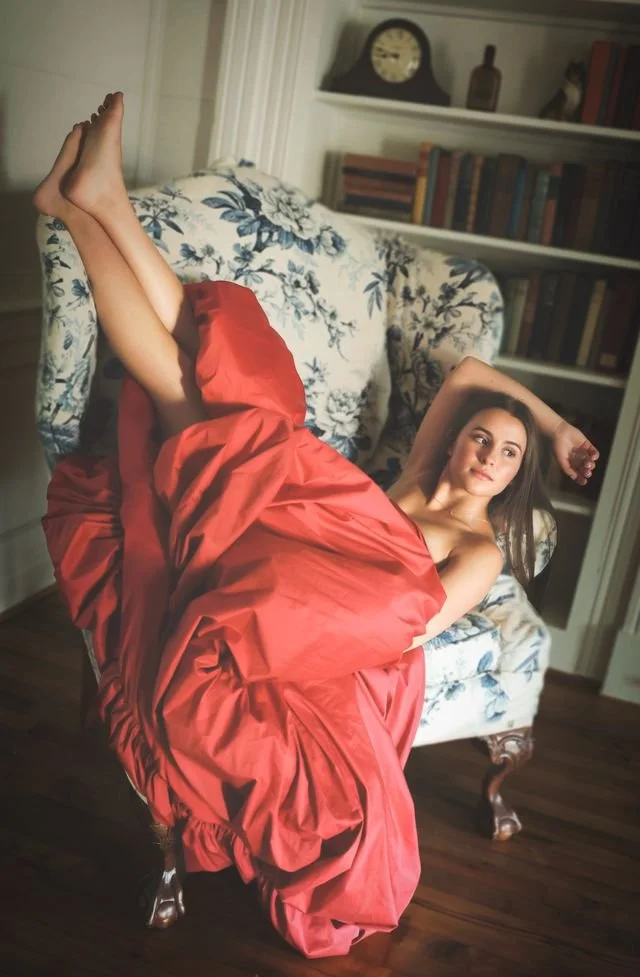 A woman in a red dress reclining on a patterned sofa with her legs raised, in a room with wooden floors and a bookshelf in the background.