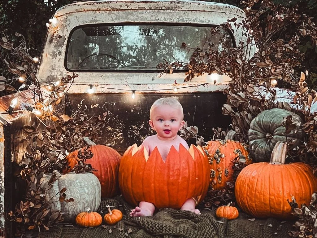 A baby is sitting inside a carved pumpkin with a surprised expression. The scene is set in the bed of an old, weathered truck decorated with pumpkins and fall foliage, with string lights adding a warm glow.