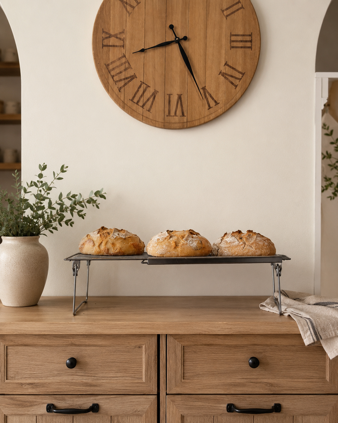 Three loaves of bread on a wire cooling rack on a wooden table, with a potted plant on the left and a clock on the wall above.