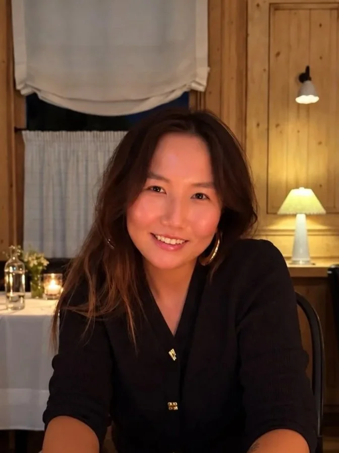 A woman with long brown hair and hoop earrings smiling at the camera in a warmly lit, cozy wooden interior with a table set for dinner in the background.