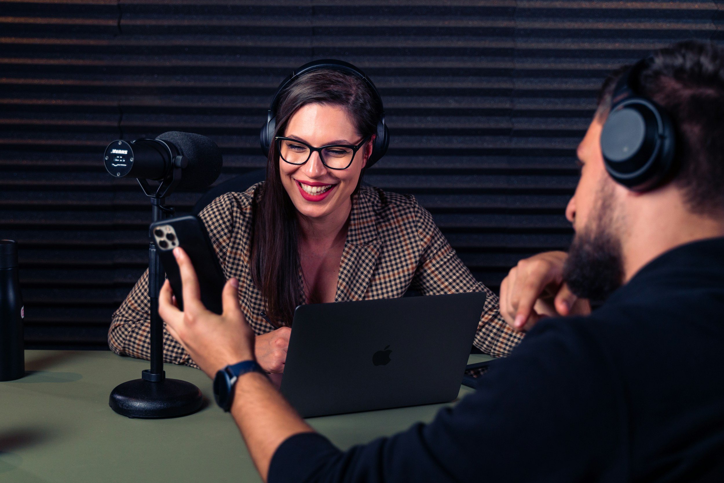 Woman with glasses smiling and holding a smartphone during a podcast recording with a man, in a studio with black acoustic foam panels.