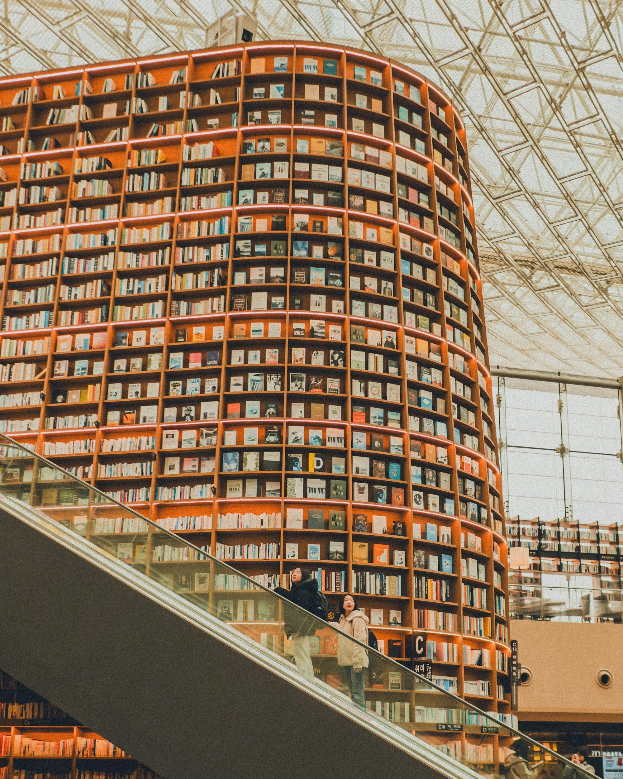 A large, round bookshelf filled with books in an indoor space, with people walking on an escalator below.