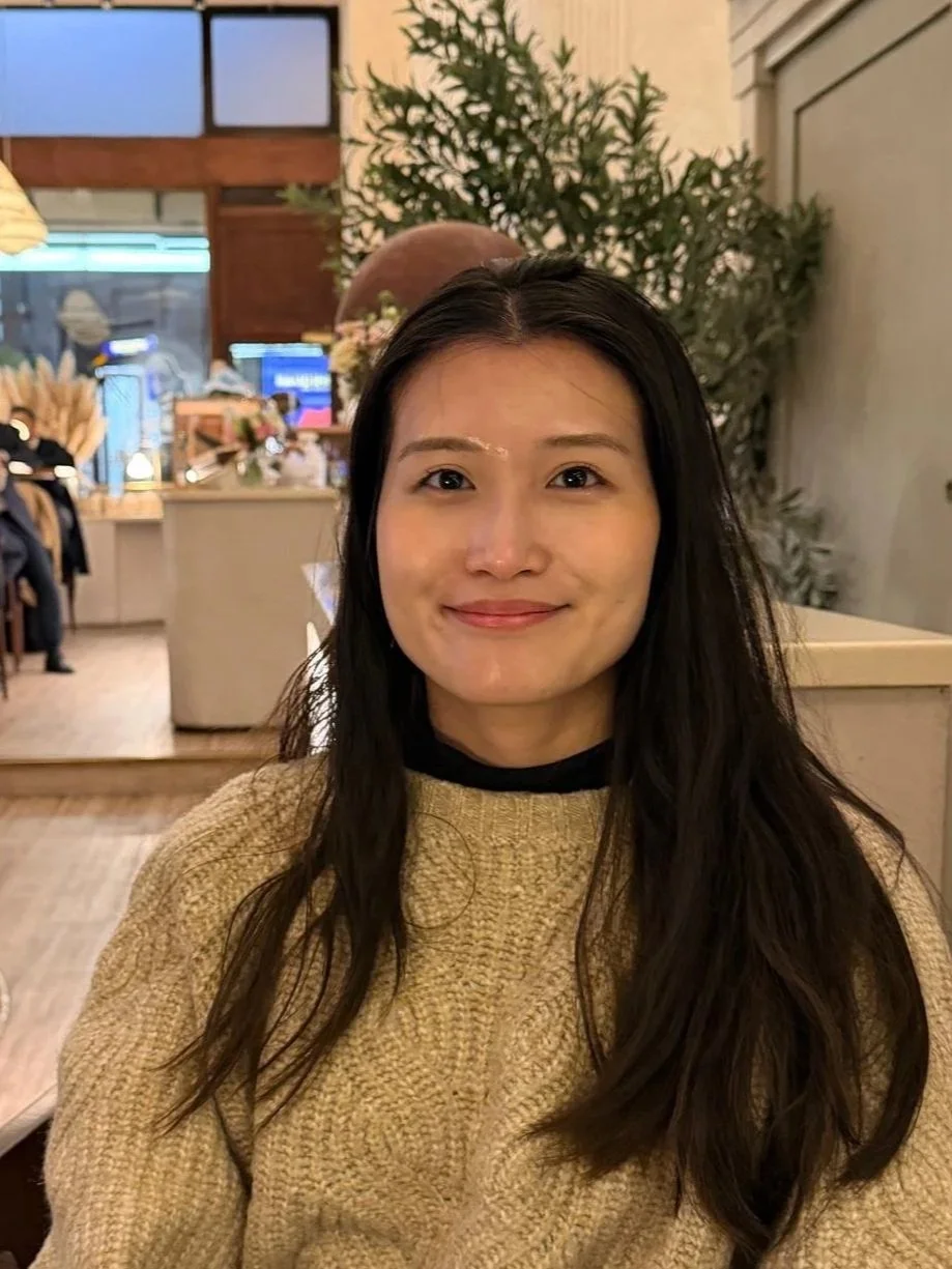 A woman with long dark hair and a beige sweater smiling at the camera inside a cozy restaurant or cafe, with plants and decorative items in the background.