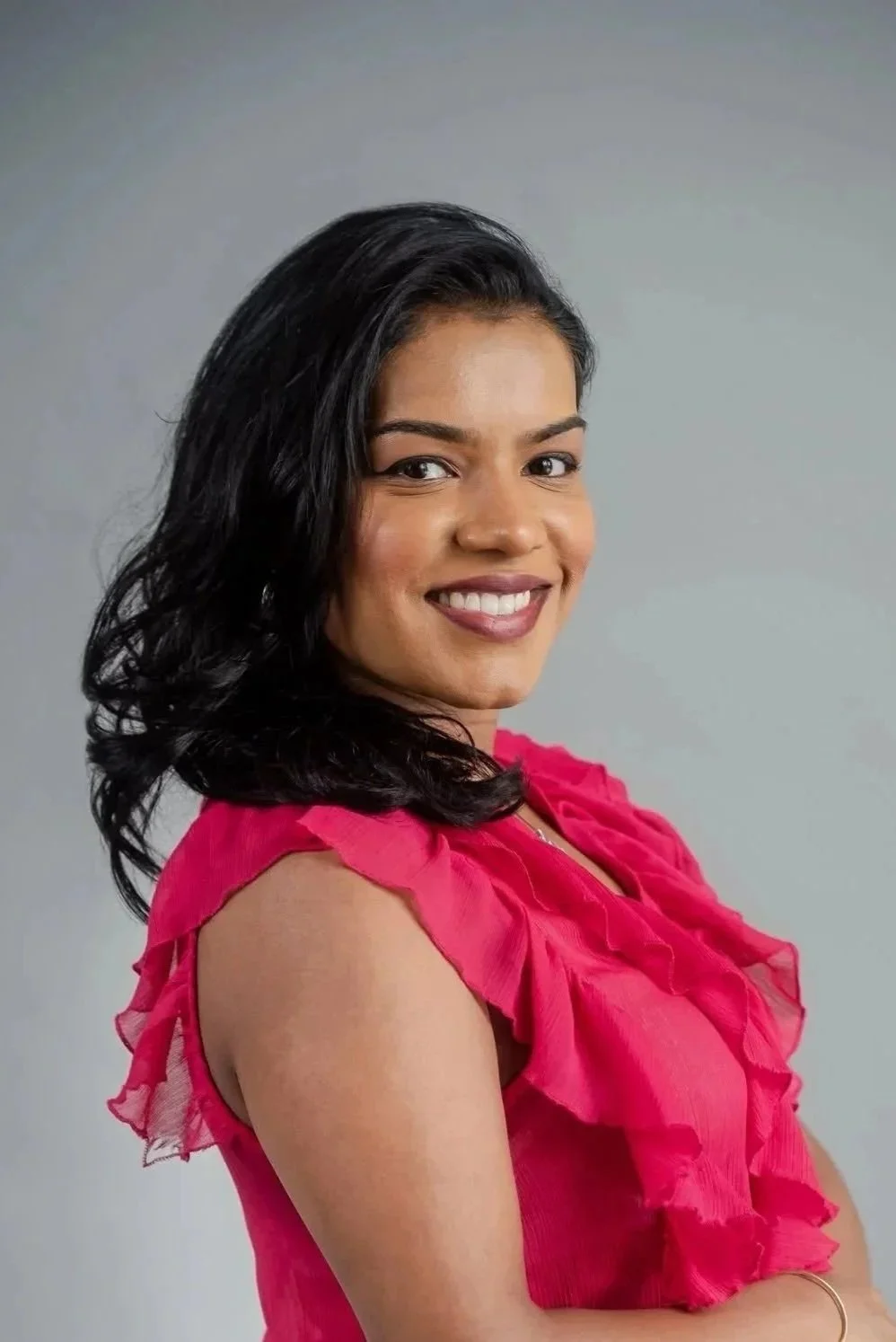 A woman with black hair, wearing a pink sleeveless blouse with ruffled details, smiling against a plain gray background.