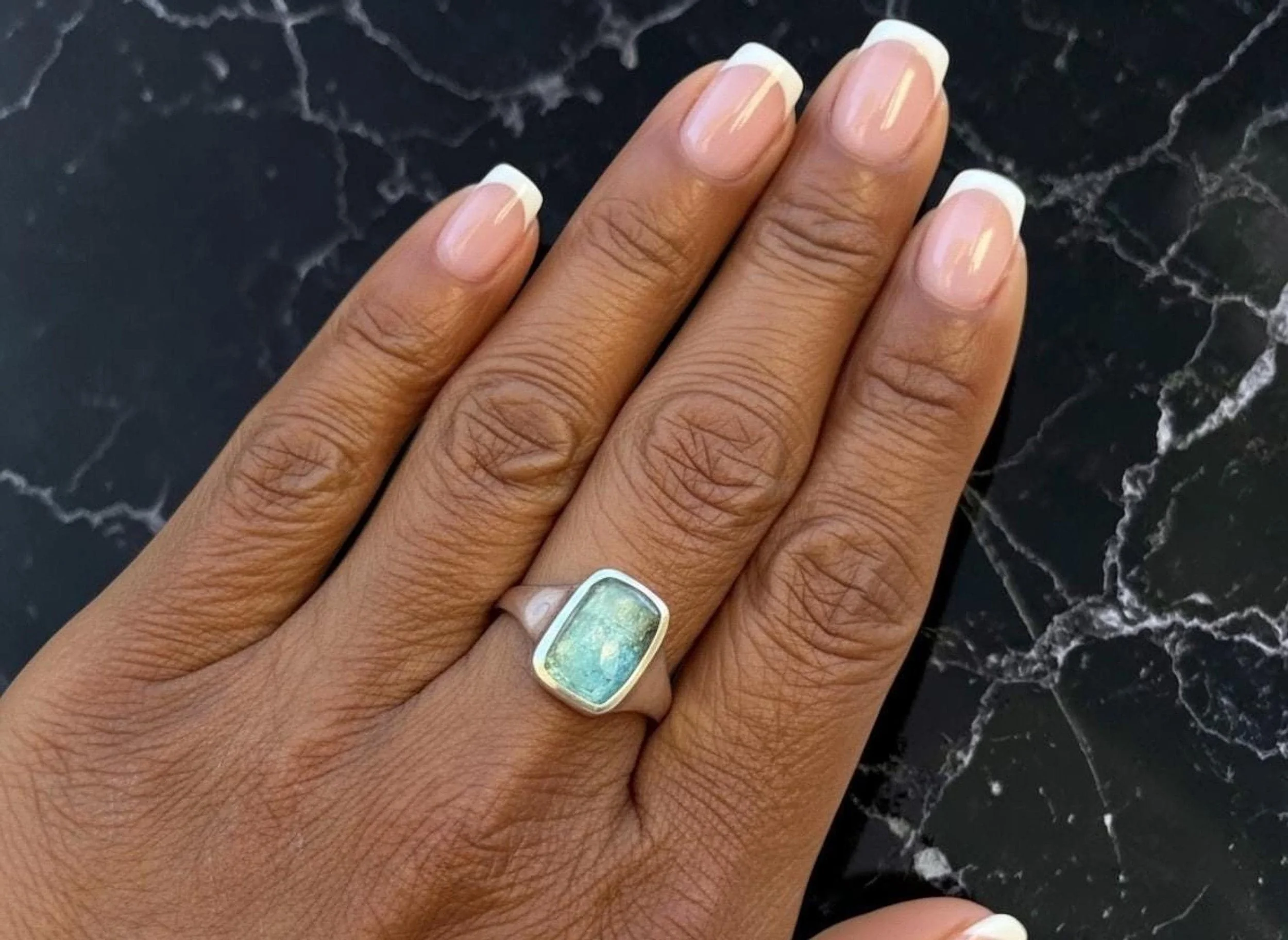Close-up of a person's hand with a nude manicure and a large, rectangular ring with a kyante stone, resting on a dark marble surface.