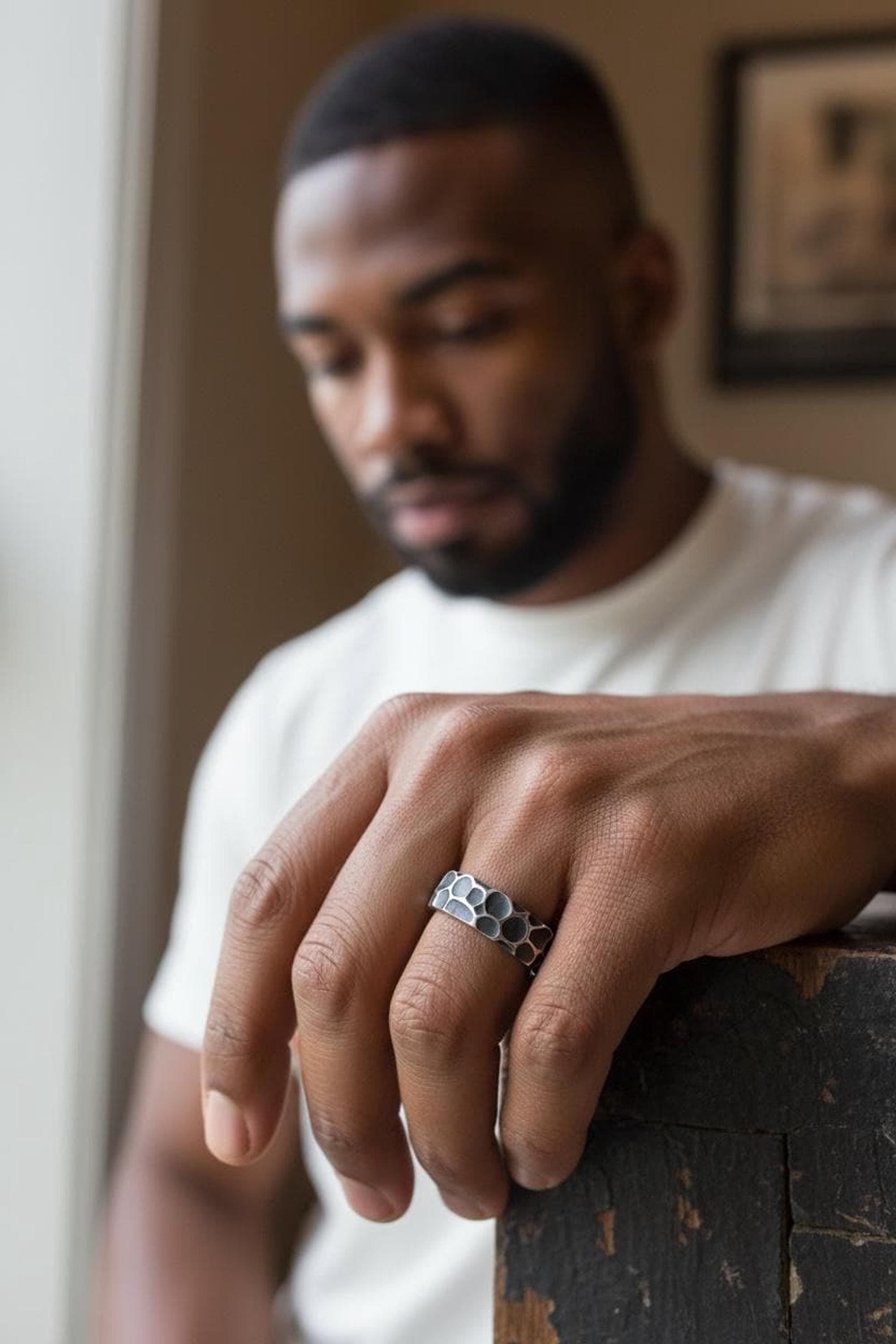 Close-up of a man's hand with a silver Honeycomb-patterned ring on his finger, resting on a dark wooden surface. The man has a beard and short hair, wearing a white shirt, with a blurred background.