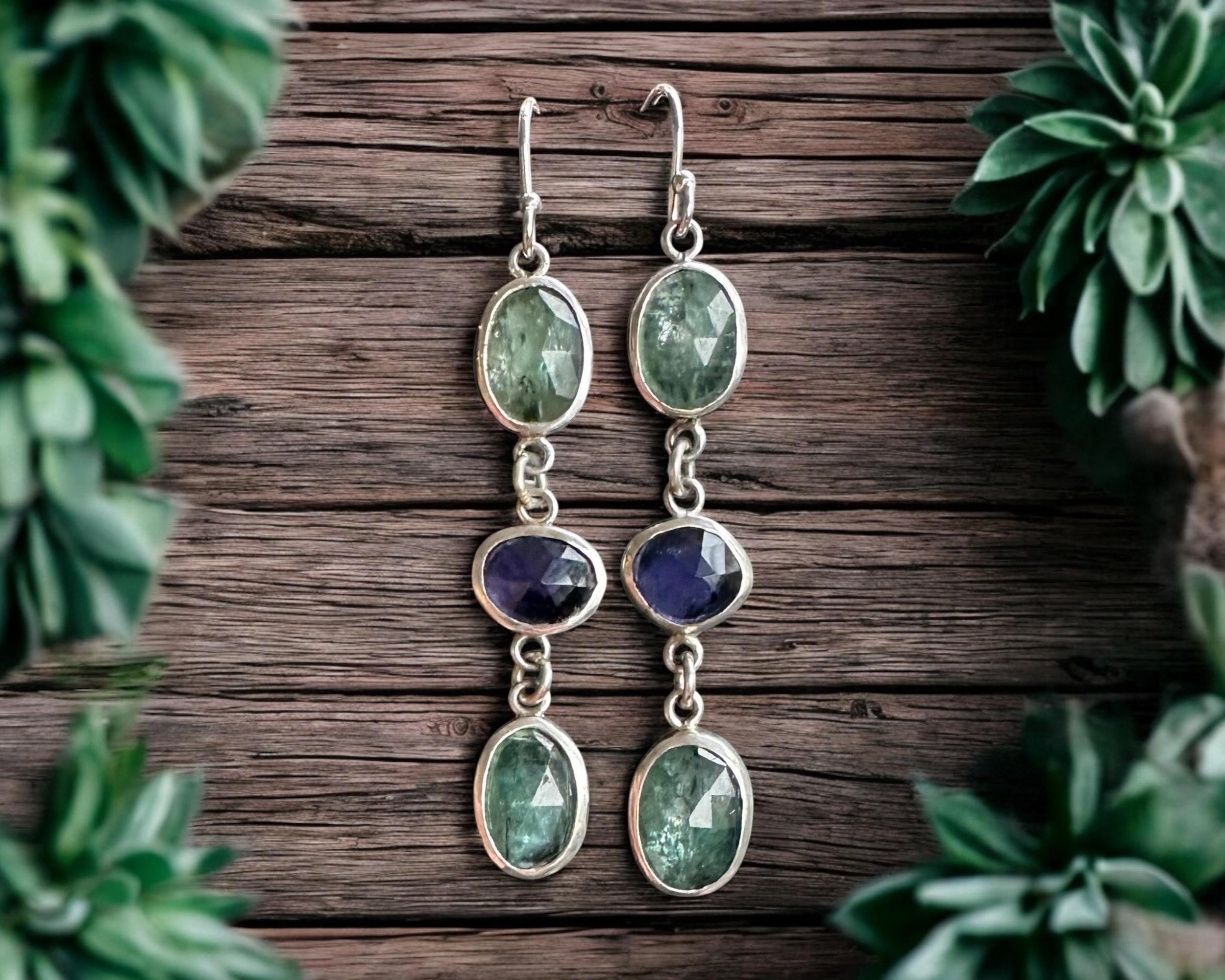 A pair of silver earrings with oval-shaped green stones hanging from silver hooks, placed on a wooden surface, with green foliage partially framing the image.