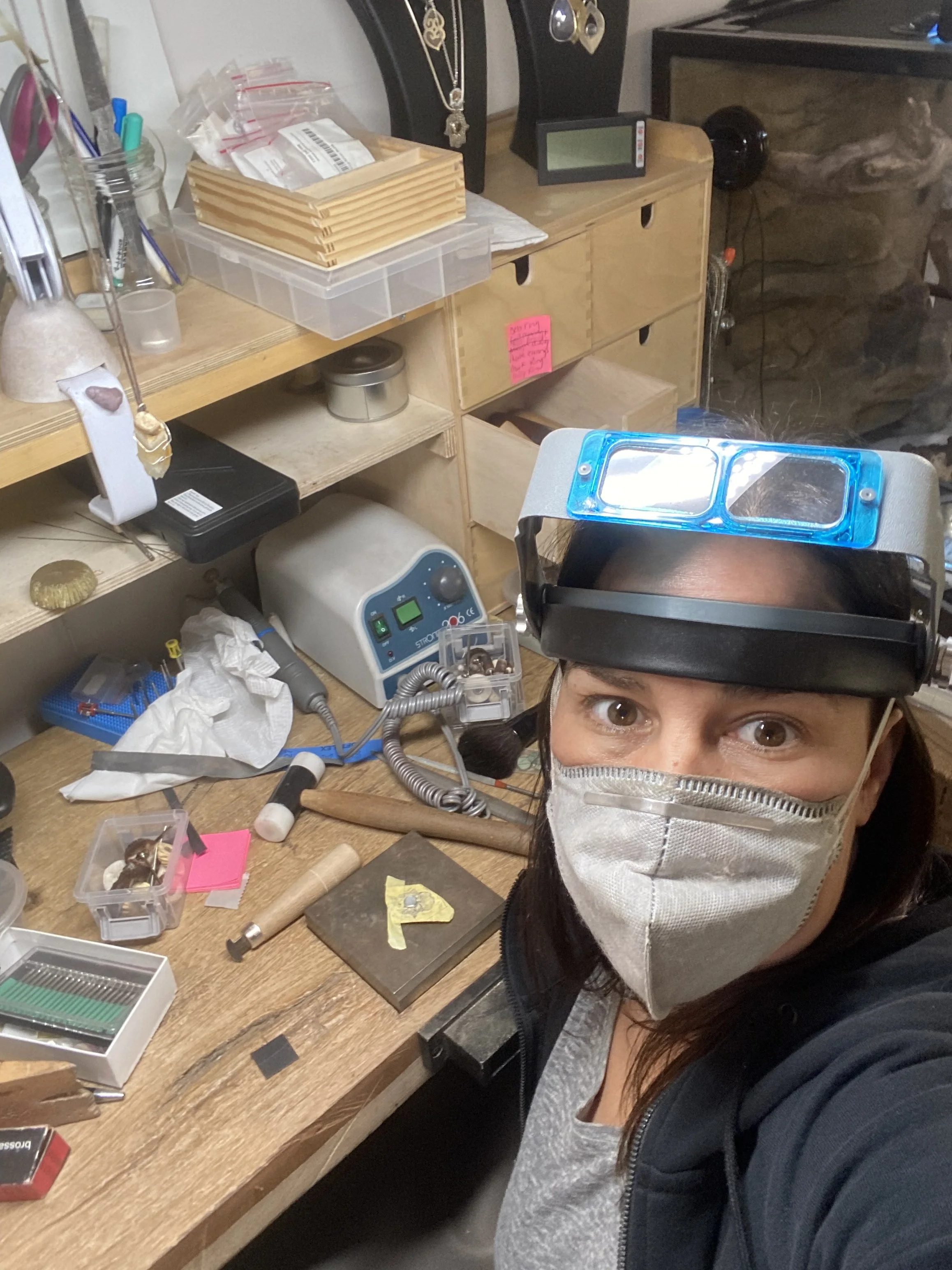 Person wearing a face mask and magnifying glasses on their forehead, working at a cluttered workshop table with various tools and equipment.