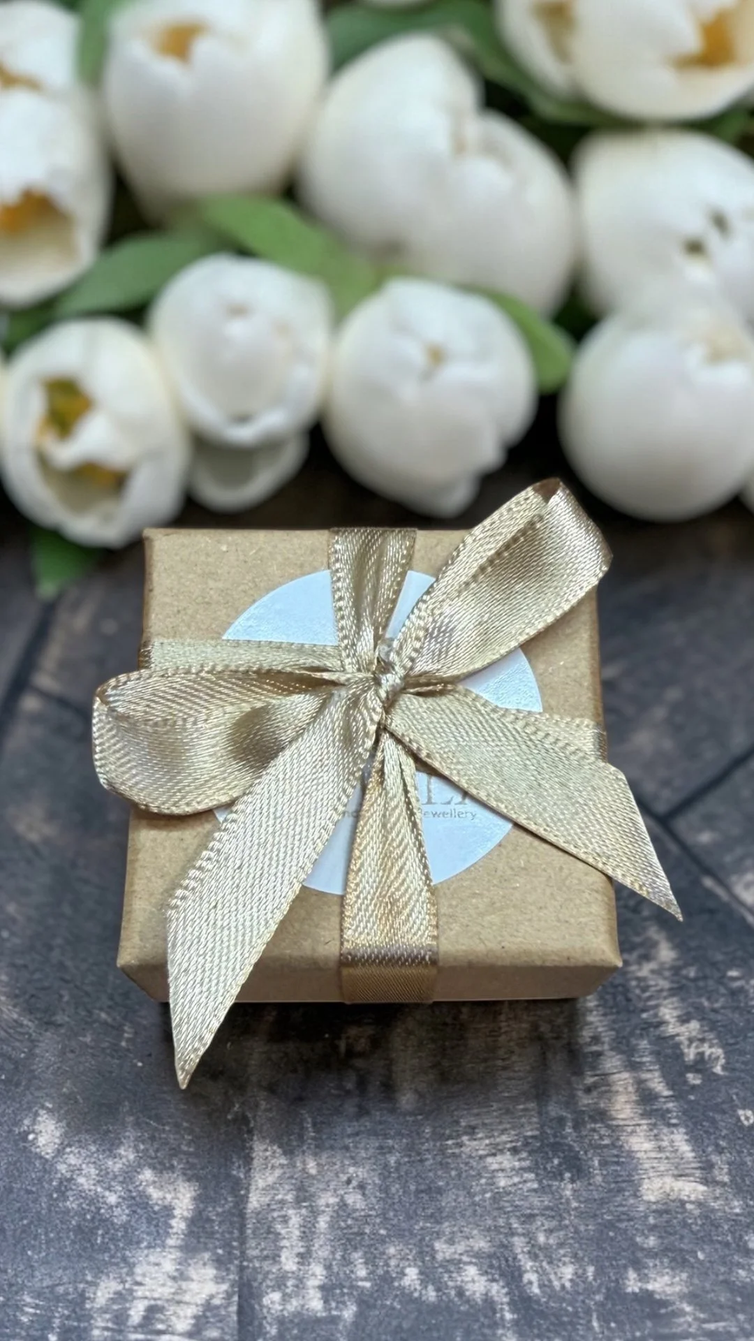 Small gift box wrapped in gold paper with a gold ribbon and bow, with white tulips in the background.