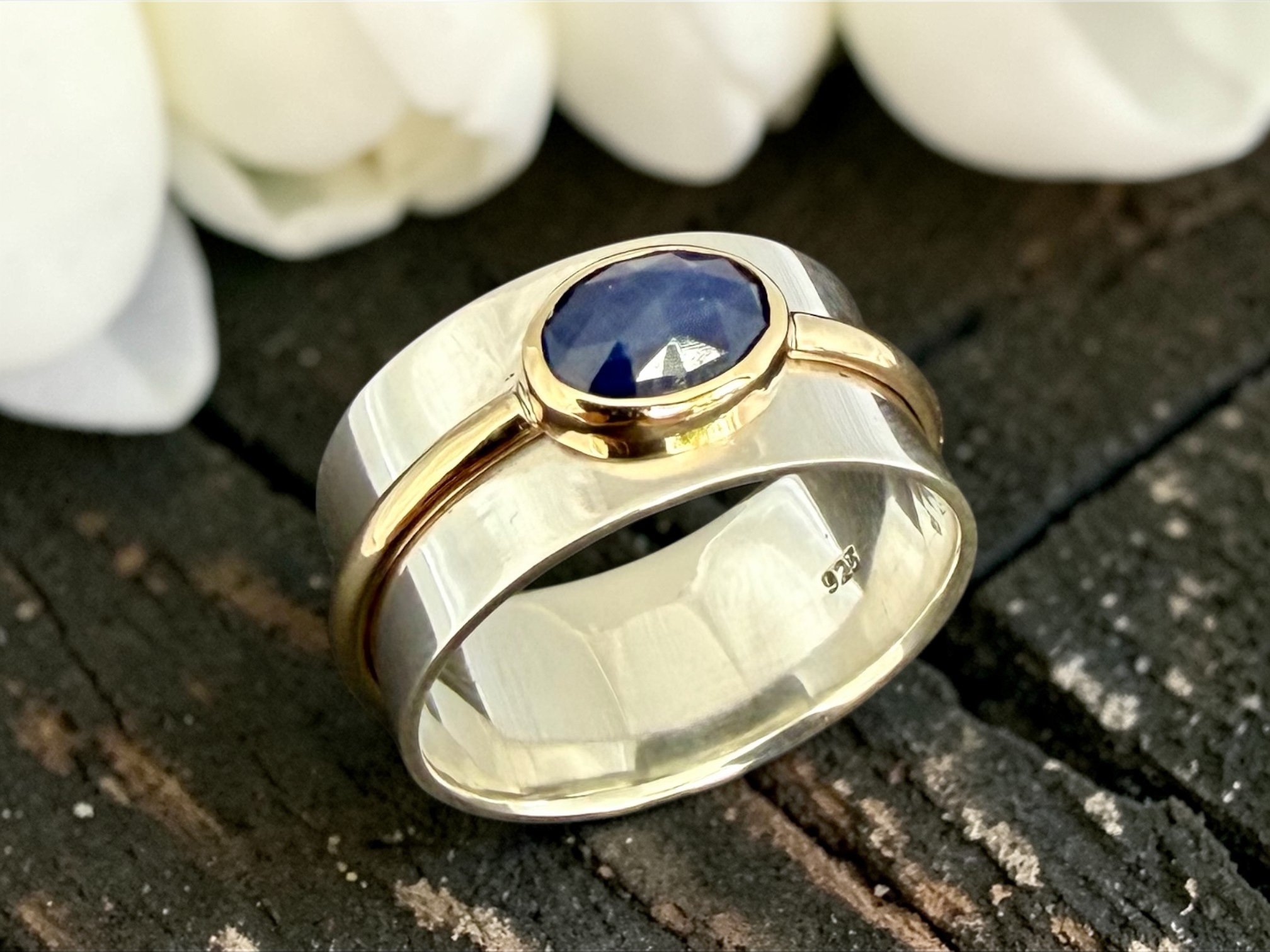 Close-up of a gold ring with a dark blue gemstone, placed on a dark textured surface with white flower petals in the background.