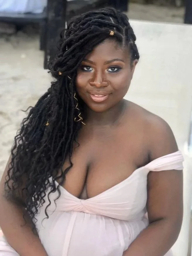 A black woman with long, styled locs and dark skin, wearing a light pink off-shoulder dress, sitting outdoors on a sandy surface.