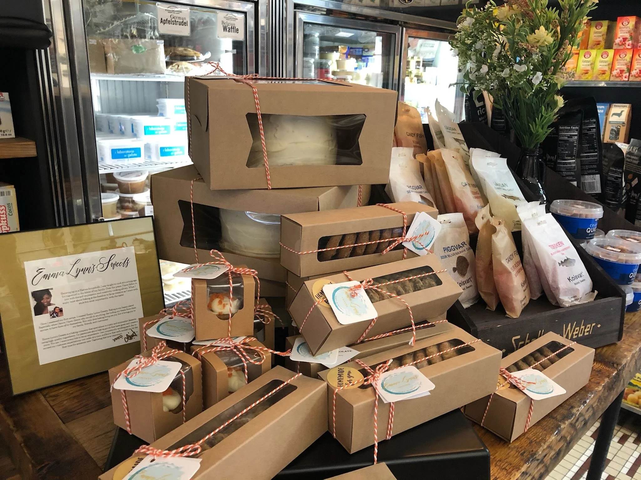 Display of packaged baked goods and treats on a counter in a shop, with flowers and refrigerated items in the background.