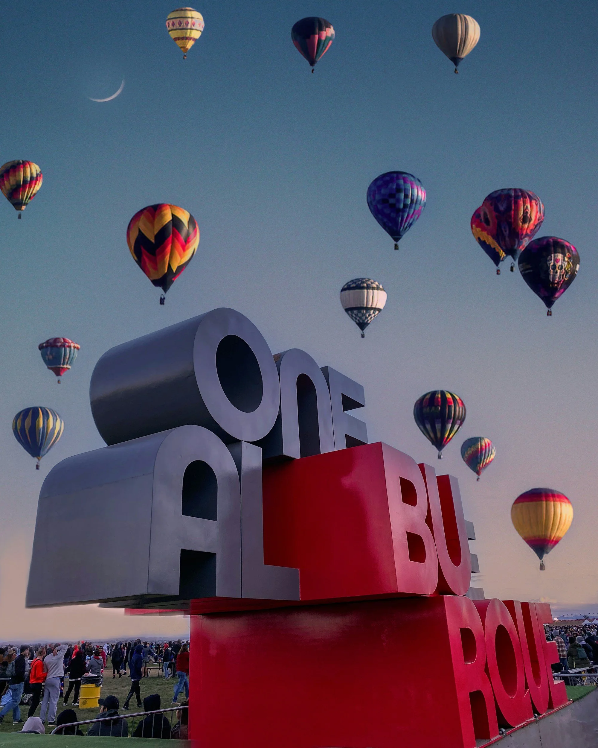 Multiple hot air balloons in various colors floating in a clear sky during an outdoor event. A large red and white sign spelling partially 'LOVE' is visible in the foreground, and a crowd of people is gathered on the ground below.