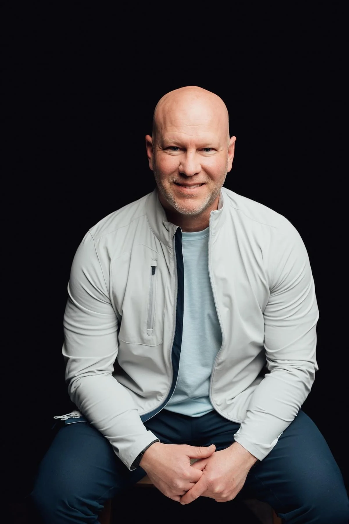 A bald man with a beard, smiling, wearing a light gray jacket over a white T-shirt, sitting against a black background.