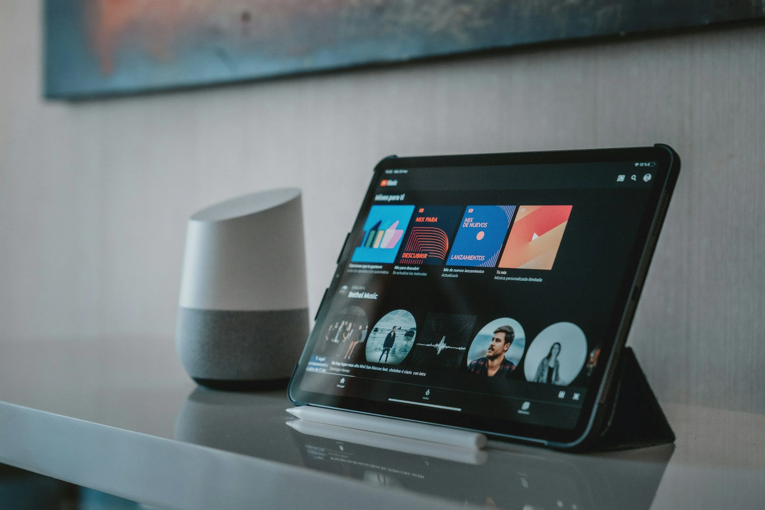 A tablet displaying a music app on a white stand, placed on a reflective white surface with a white speaker and a painting hanging on a gray wall in the background.