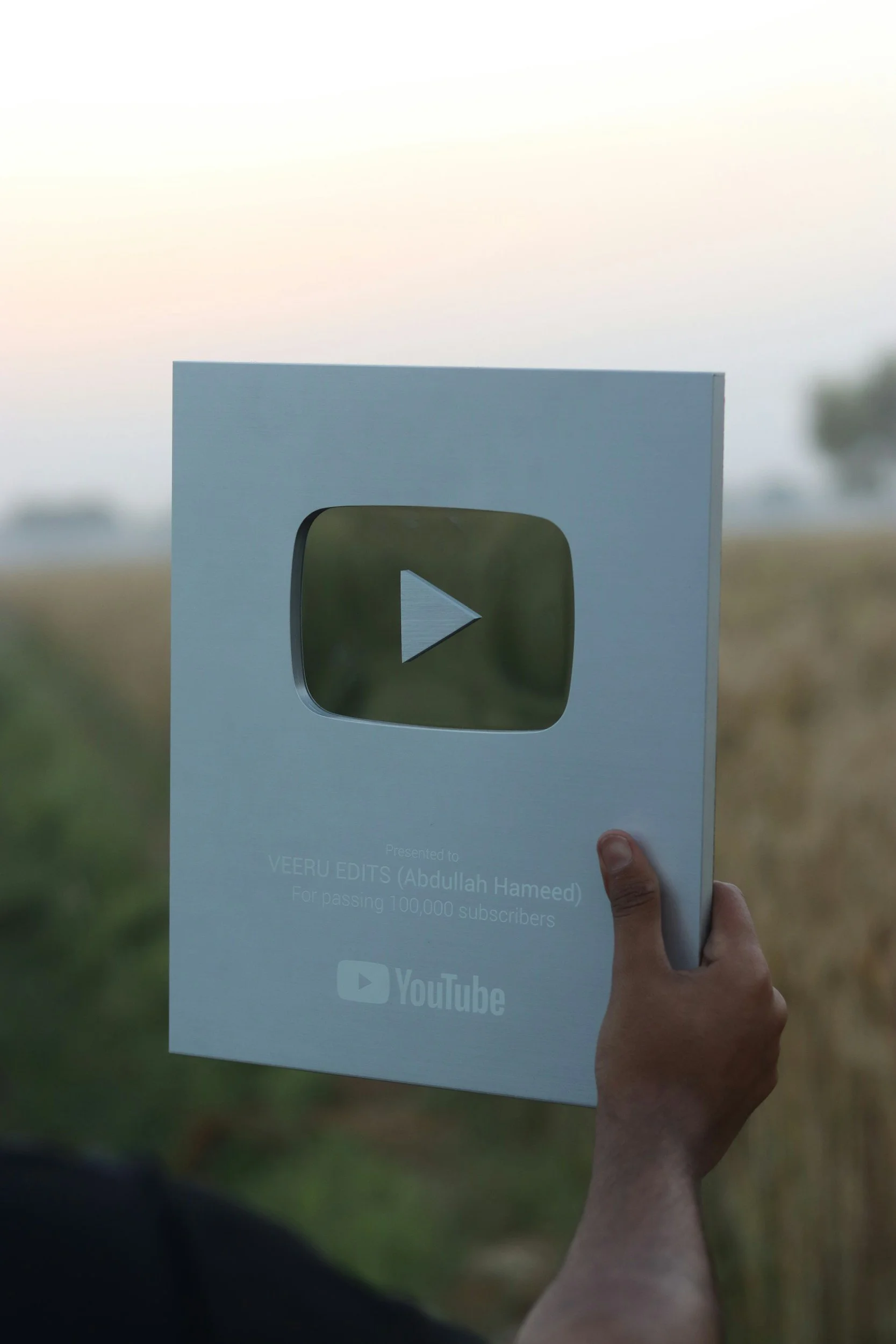 Person holding a YouTube plaque award outdoors. The plaque features a play button icon and text recognizing Abdulllah Hameed for passing 100,000 subscribers.