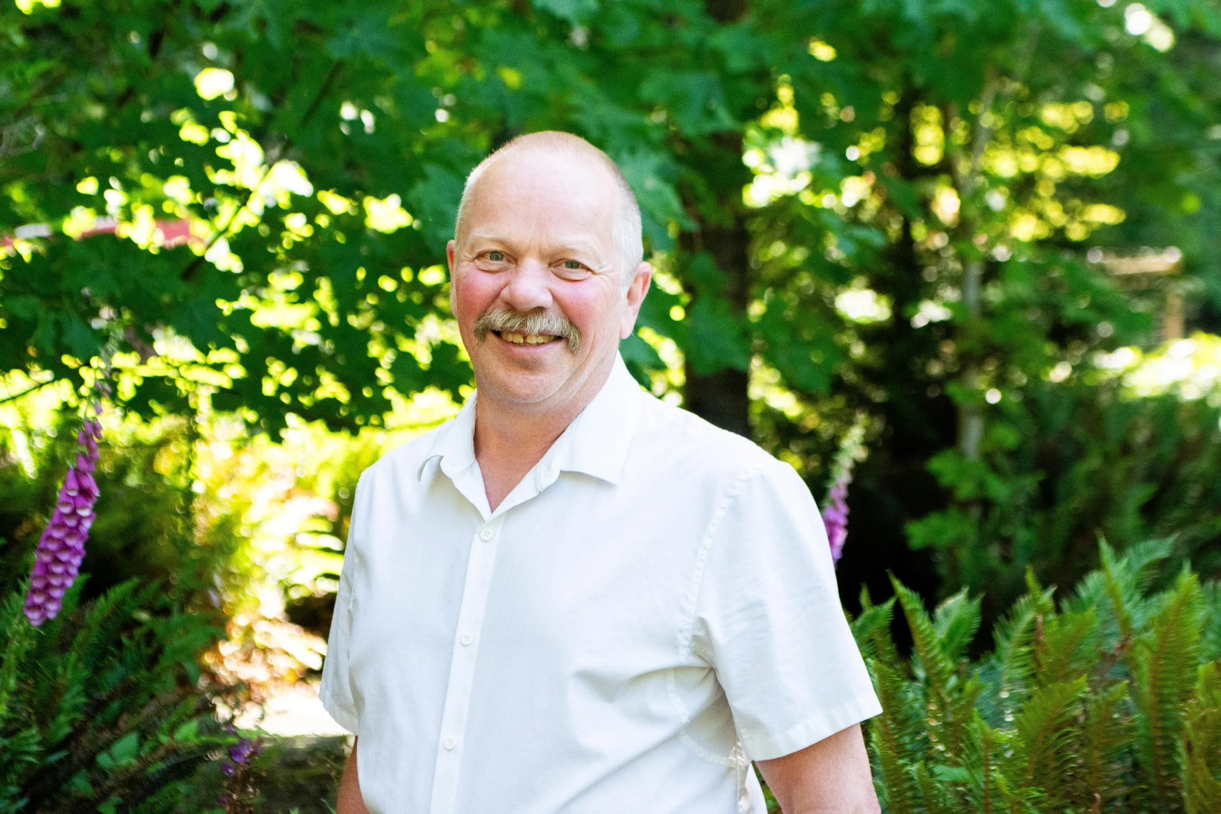 A middle-aged man with a mustache and short hair smiling outdoors in a lush, green garden, wearing a white short-sleeved shirt.