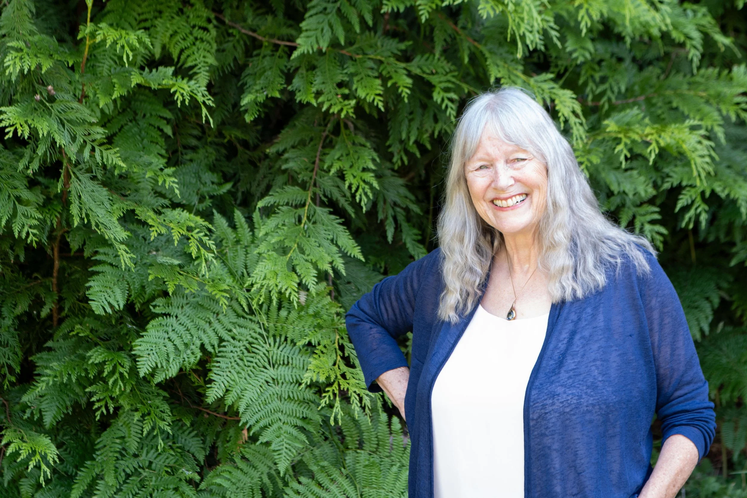 Smiling elderly woman with long gray hair standing outdoors in front of lush green ferns, wearing a white shirt and a blue jacket.