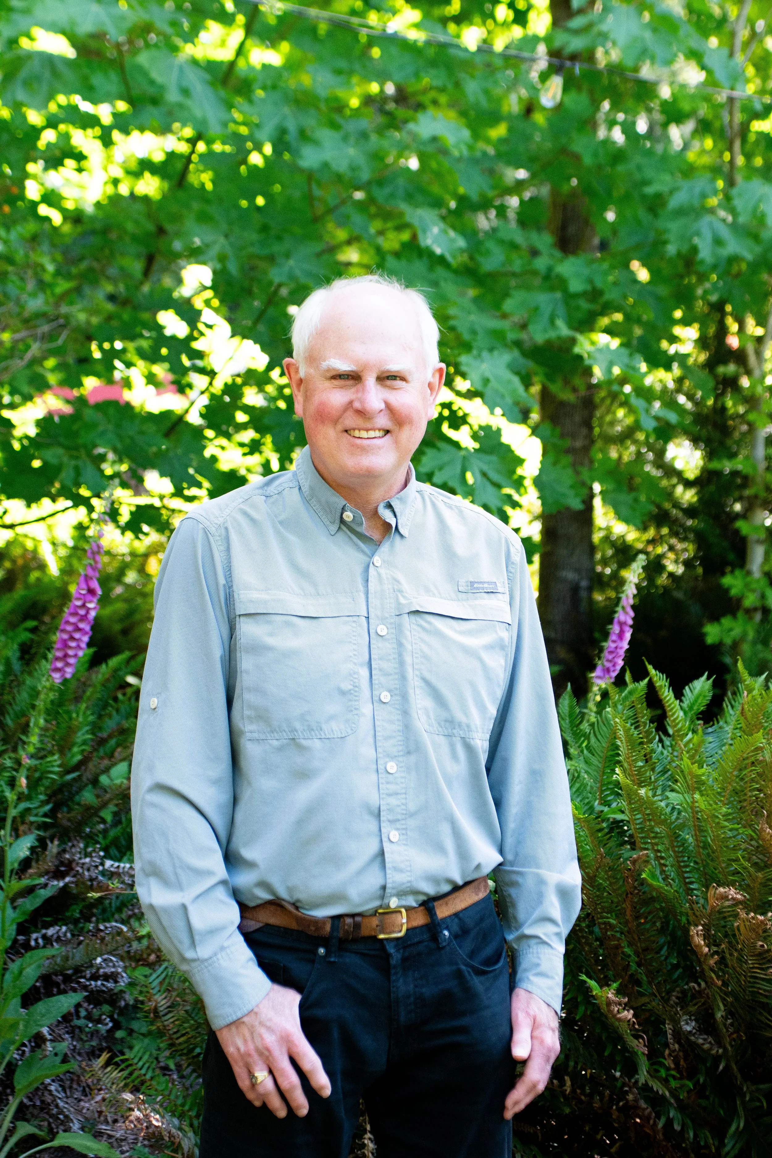Older man with white hair and a light gray button-up shirt smiling outdoors, surrounded by green foliage and purple flowers.