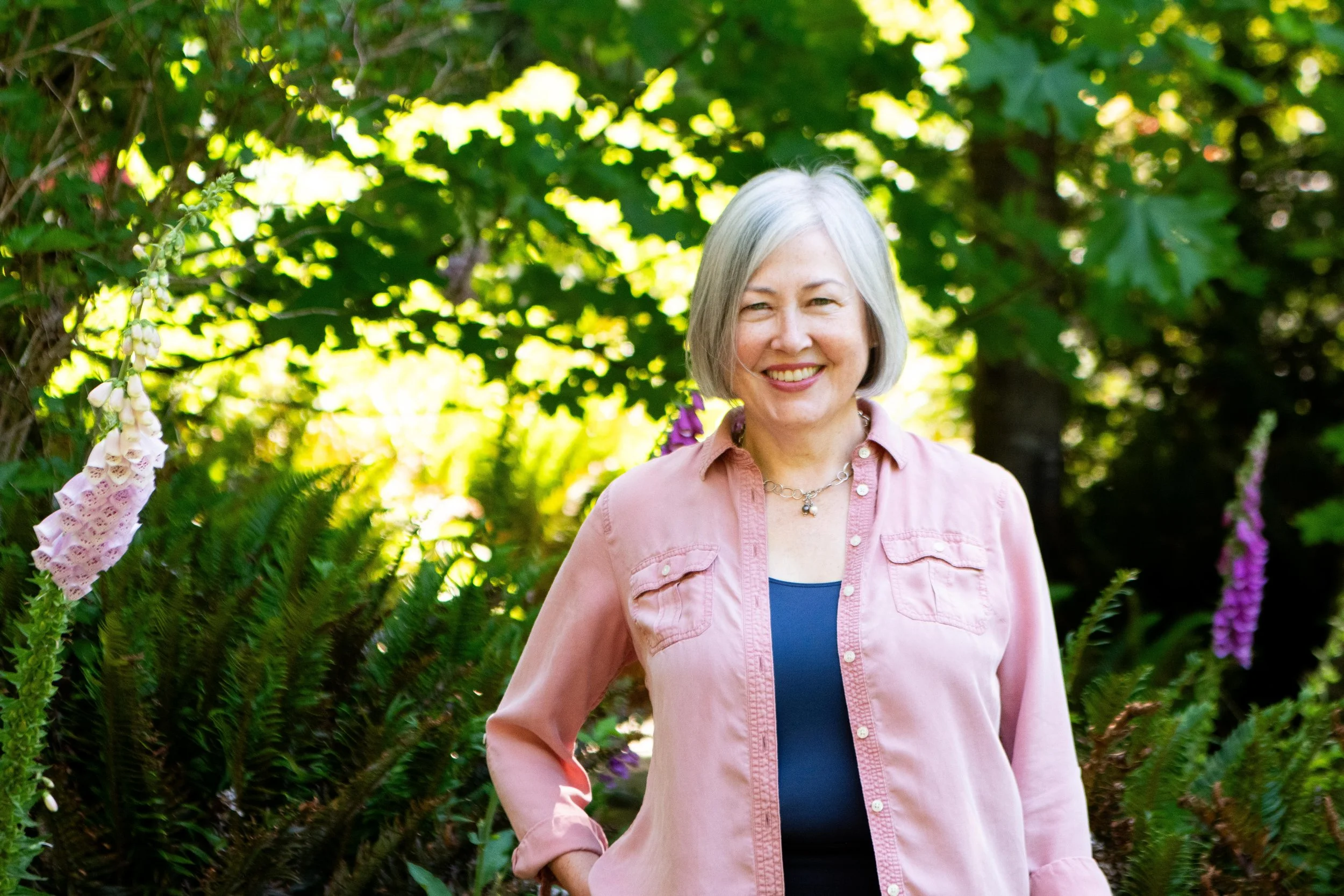 A smiling woman with gray hair standing outdoors among green foliage and purple flowers.