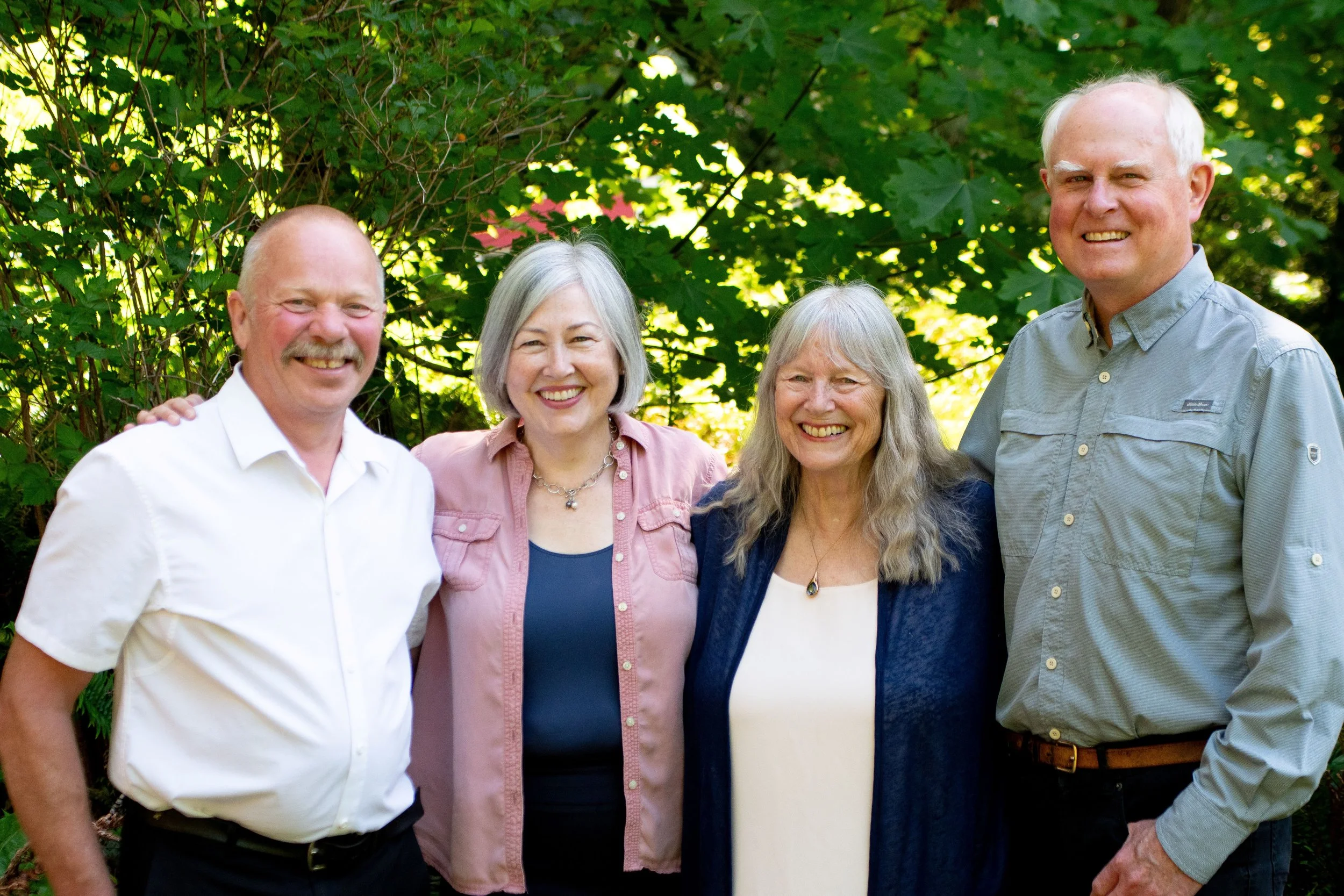 Group of five smiling adults standing outdoors in front of green leafy trees.