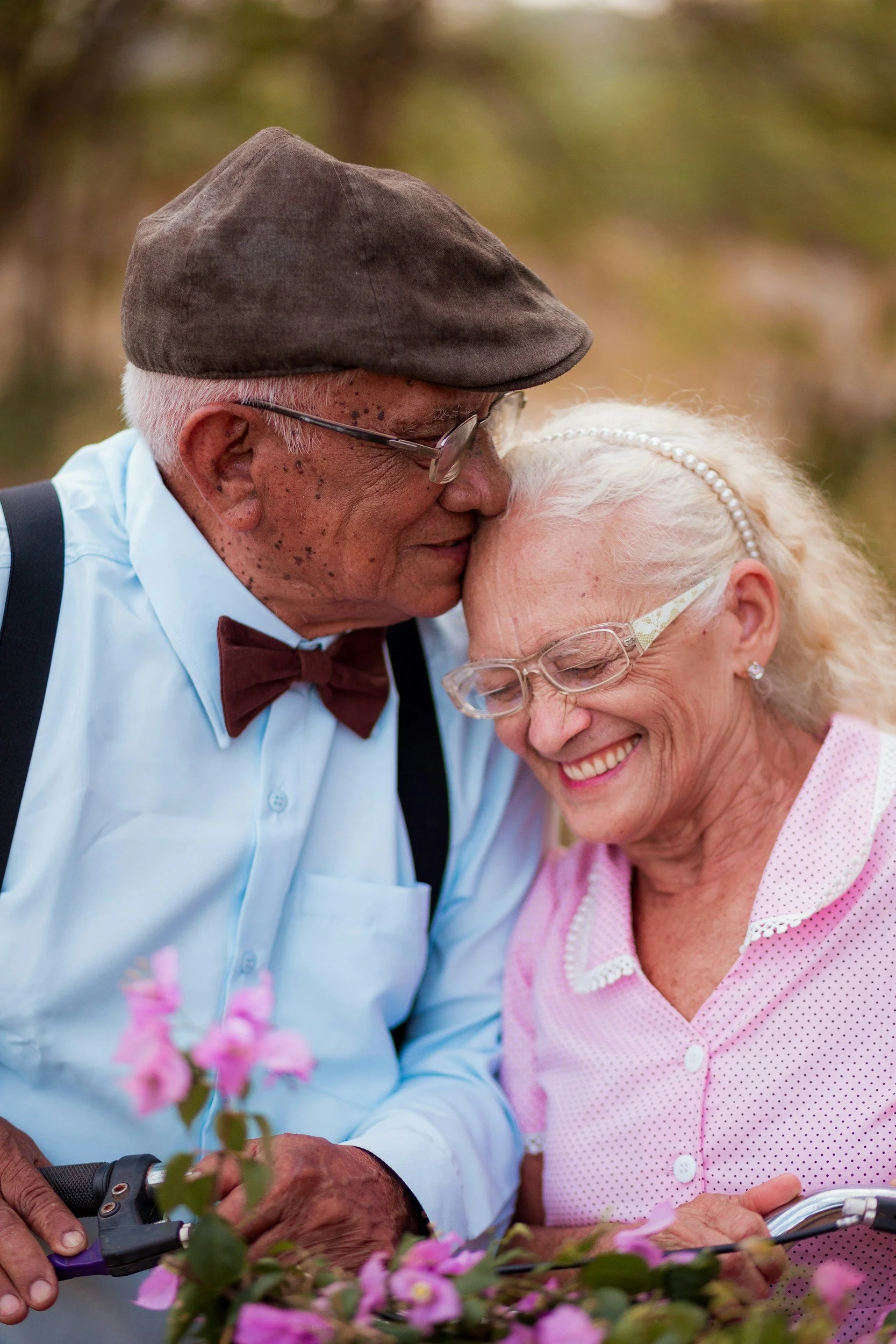 An elderly man and woman share a tender moment, close and smiling with their foreheads touching, amidst pink flowers, possibly in a garden.