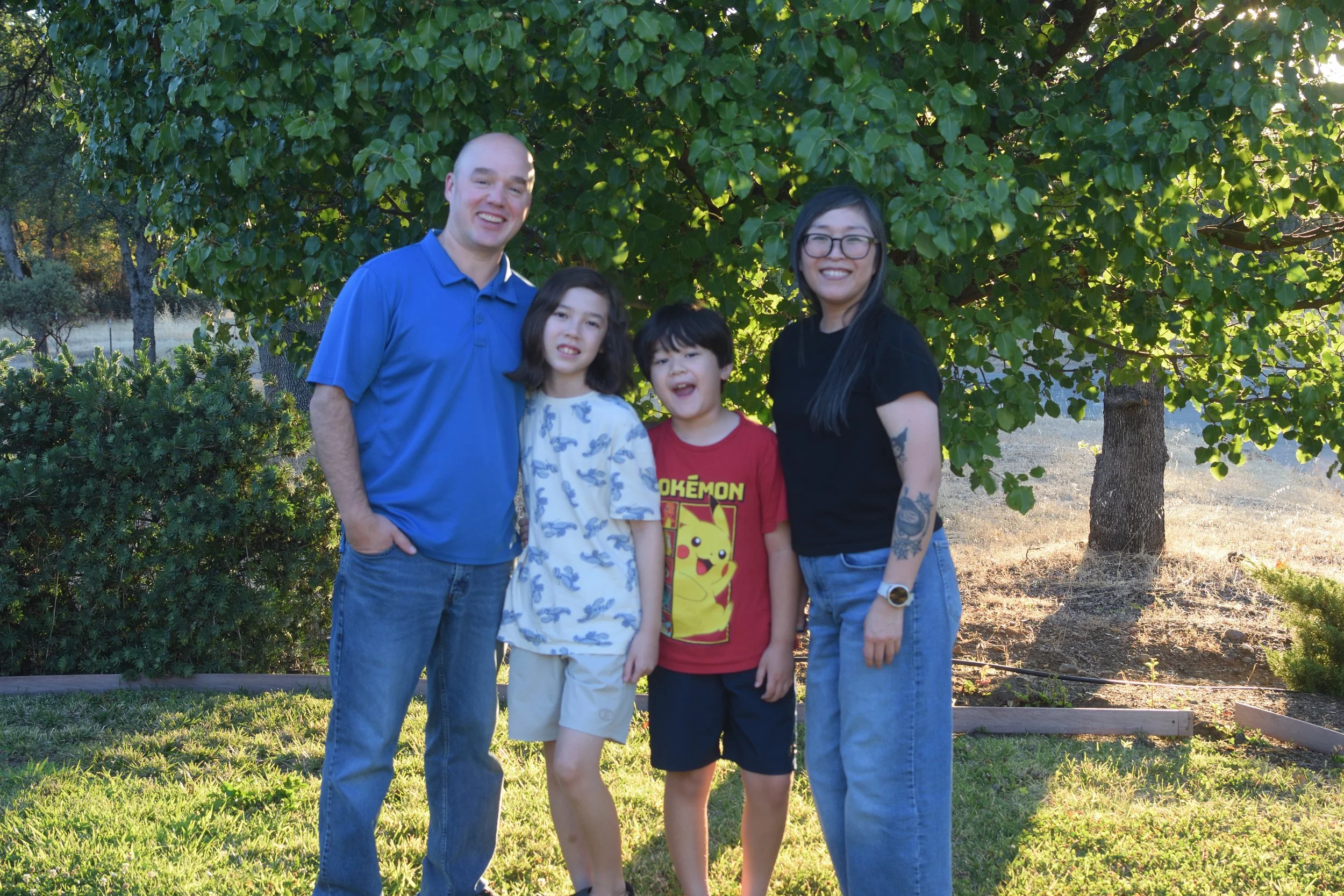 A family of five standing outdoors in front of a large leafy tree, smiling and posing for the camera.