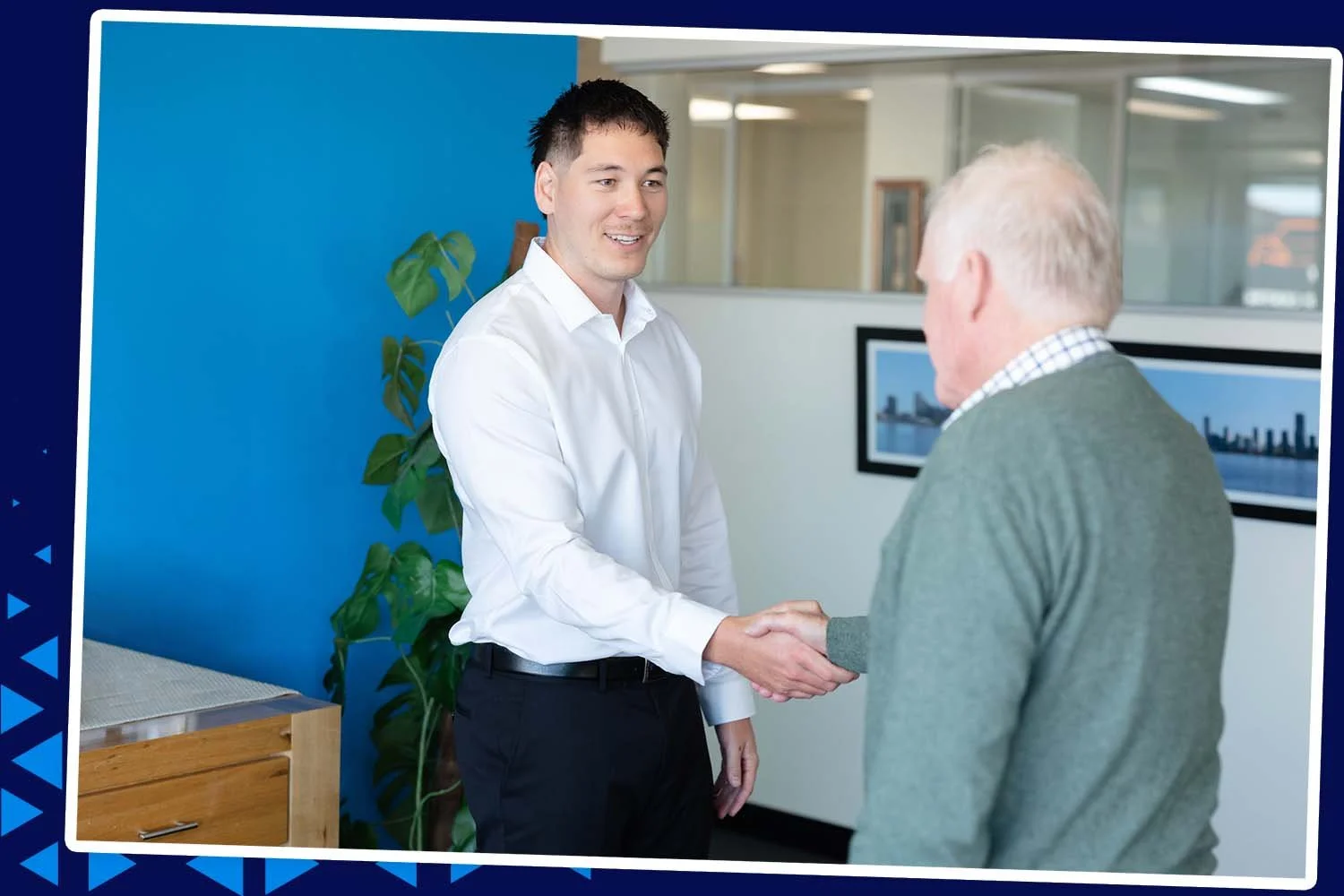 A young man in a white shirt shaking hands with an older man in a green sweater in an office setting.