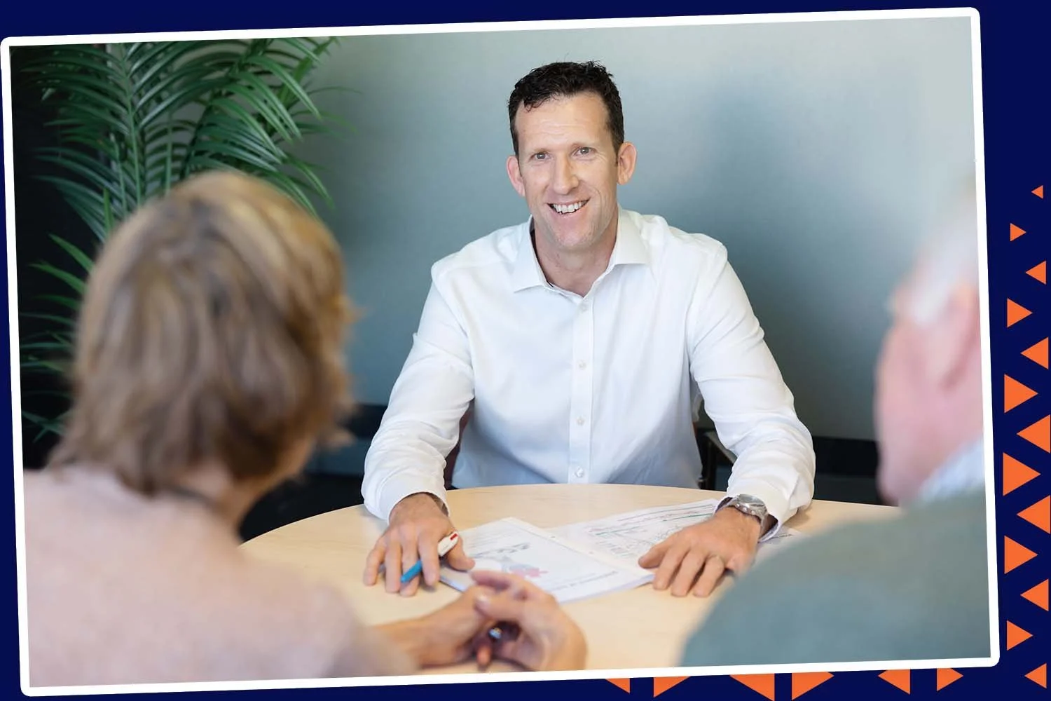 A man in a white shirt smiling during a meeting with two other people at a round table, with documents and a pen in front of him.